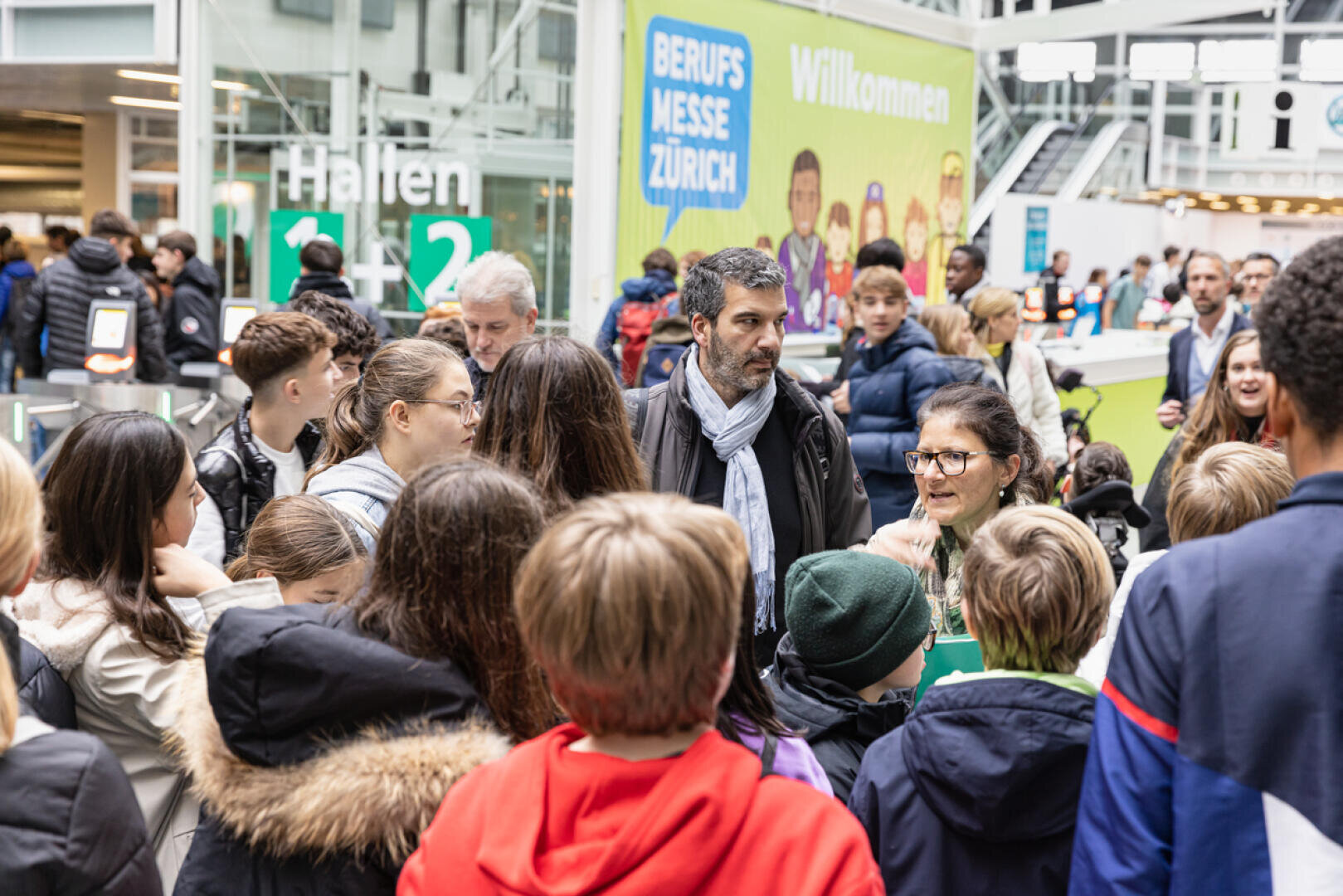 Eine Gruppe von Menschen, darunter Jugendliche und Erwachsene, versammelt sich in einem Innenraum bei einer geschäftigen Veranstaltung. Im Hintergrund weisen Schilder mit der Aufschrift "Berufs Messe Zürich" und "Willkommen" auf eine Karrieremesse in Zürich hin.
