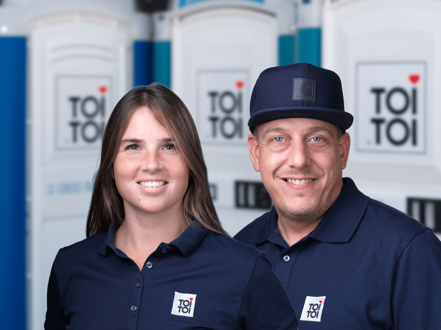 Ein Mann und eine Frau in identischen marineblauen Poloshirts mit dem TOI TOI-Logo, die vor mobilen Toiletten stehen.