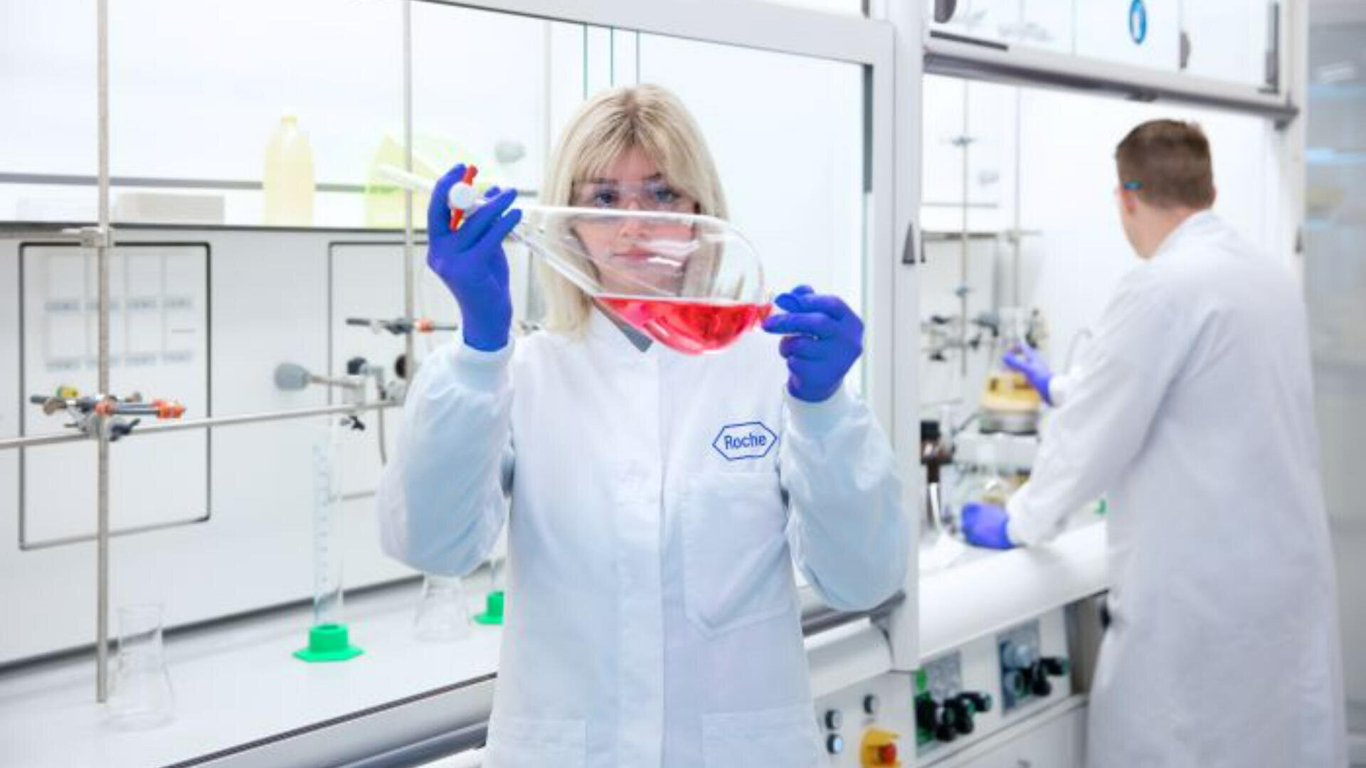 Ein Wissenschaftler in einem weißen Laborkittel und mit blauen Handschuhen hält einen großen Kolben mit roter Flüssigkeit in einem Labor, während eine andere Person im Hintergrund arbeitet, umgeben von Laborgeräten und Glaswaren.