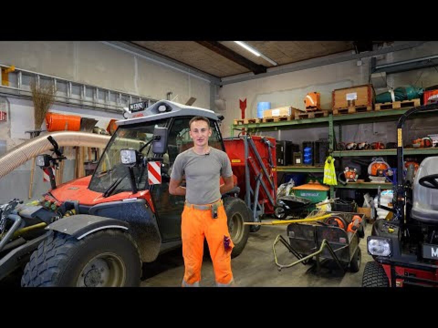 Ein Mann in einer orangefarbenen Arbeitshose und einem grauen Hemd steht in einer Garage oder Werkstatt, umgeben von Nutzfahrzeugen, Werkzeugen und Regalen mit Geräten und Zubehör.
