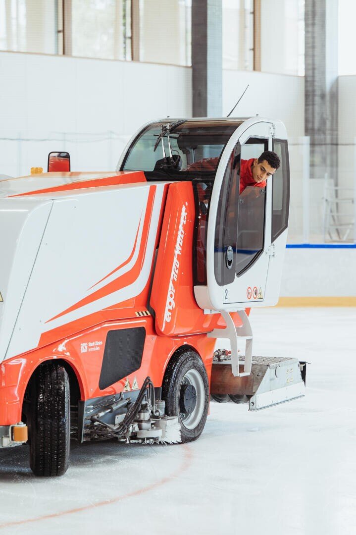 Eine Person, die eine leuchtend orange-weiße Eisbearbeitungsmaschine auf einer überdachten Eisbahn fährt, schaut aus der offenen Kabinentür.