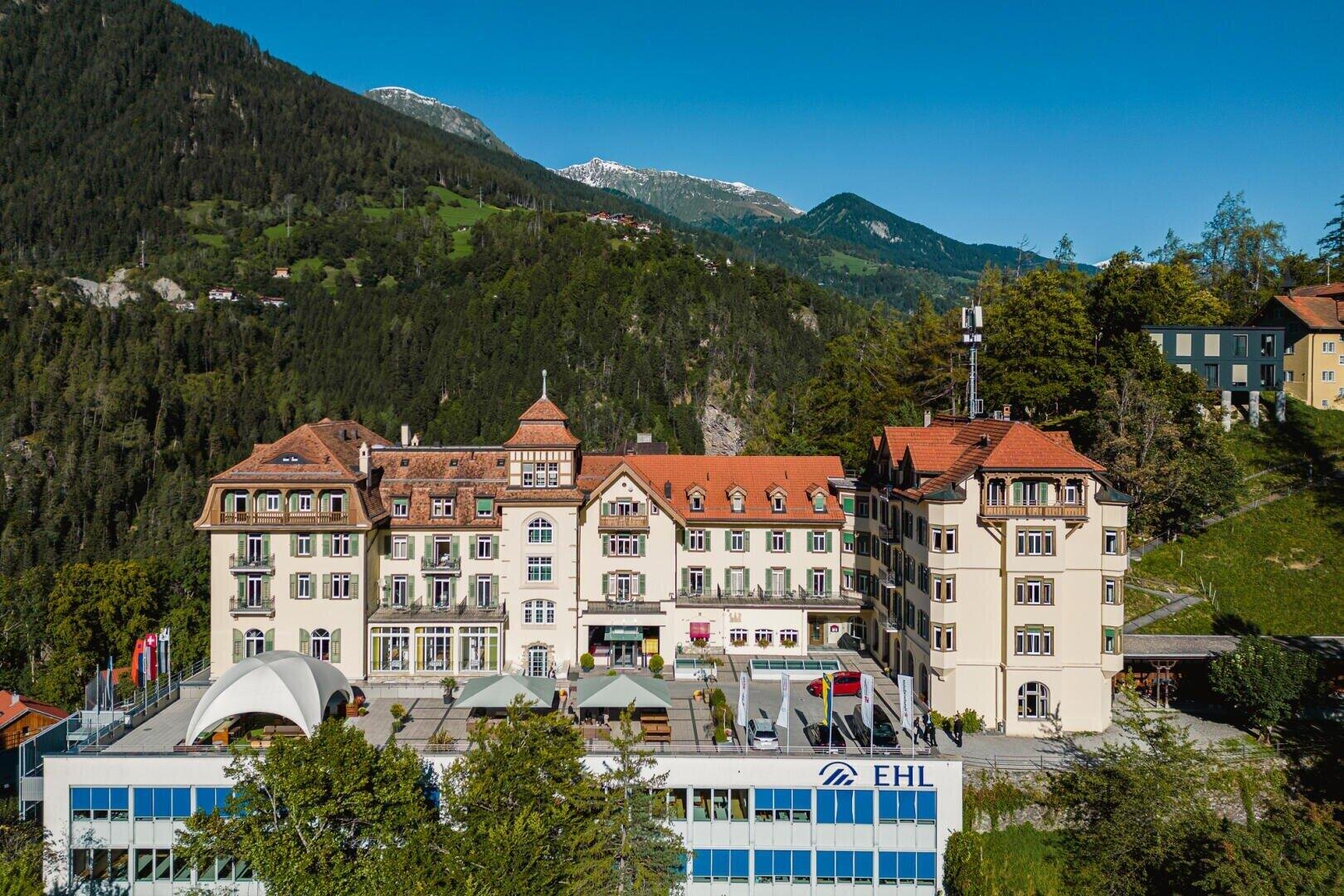 Ein großes Gebäude mit roten Dächern und klassischer Architektur steht inmitten grüner Hügel und Berge unter einem strahlend blauen Himmel. Davor ist ein modernes Gebäude mit einem blauen EHL-Schild zu sehen. Die Gebäude sind von Bäumen umgeben.