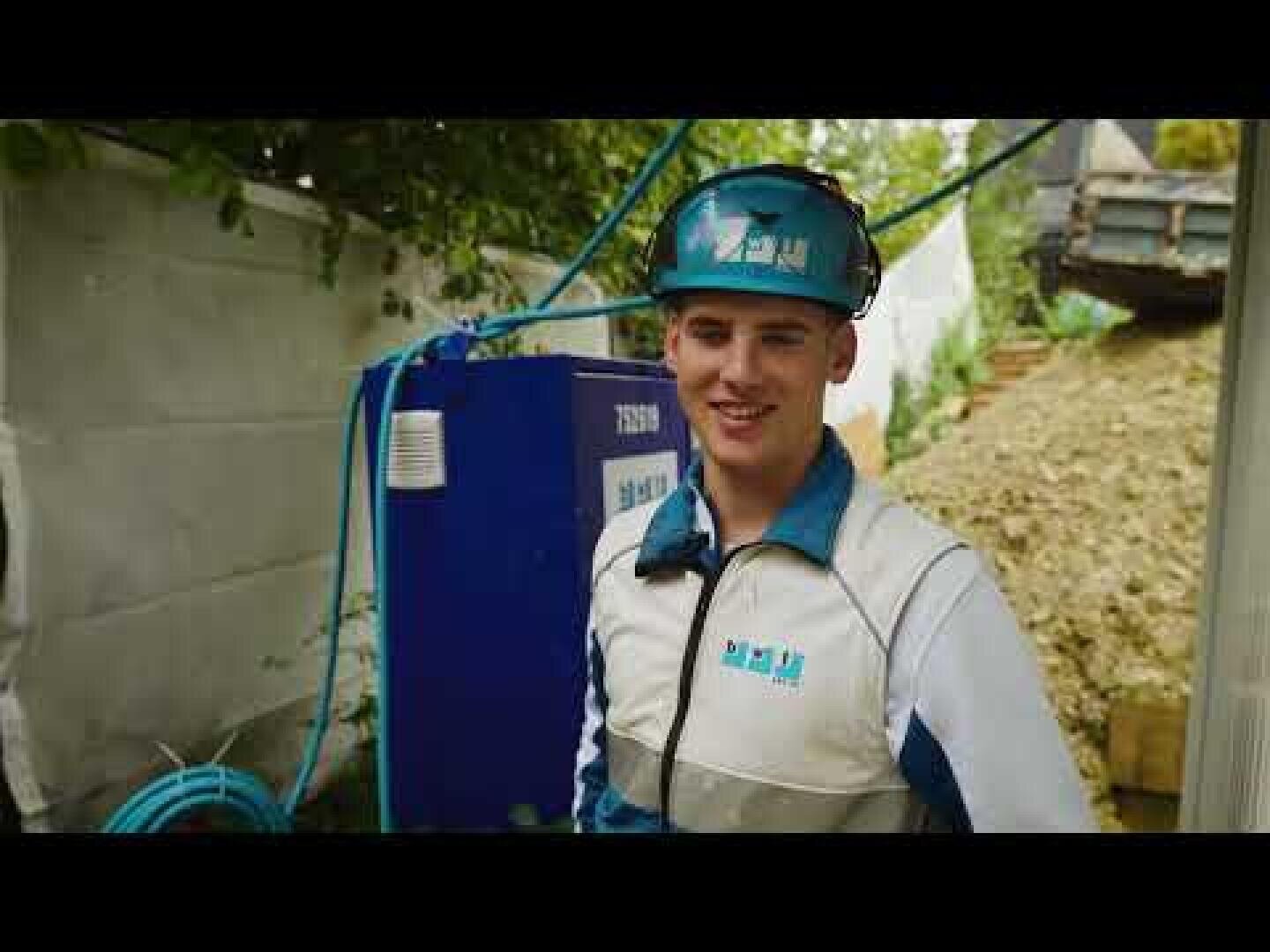 Ein lächelnder Bauarbeiter, der einen blauen Helm und eine weiß-blaue Uniform trägt, steht im Freien in der Nähe von Geräten und Kabeln, mit Bäumen und einem Zaun im Hintergrund.