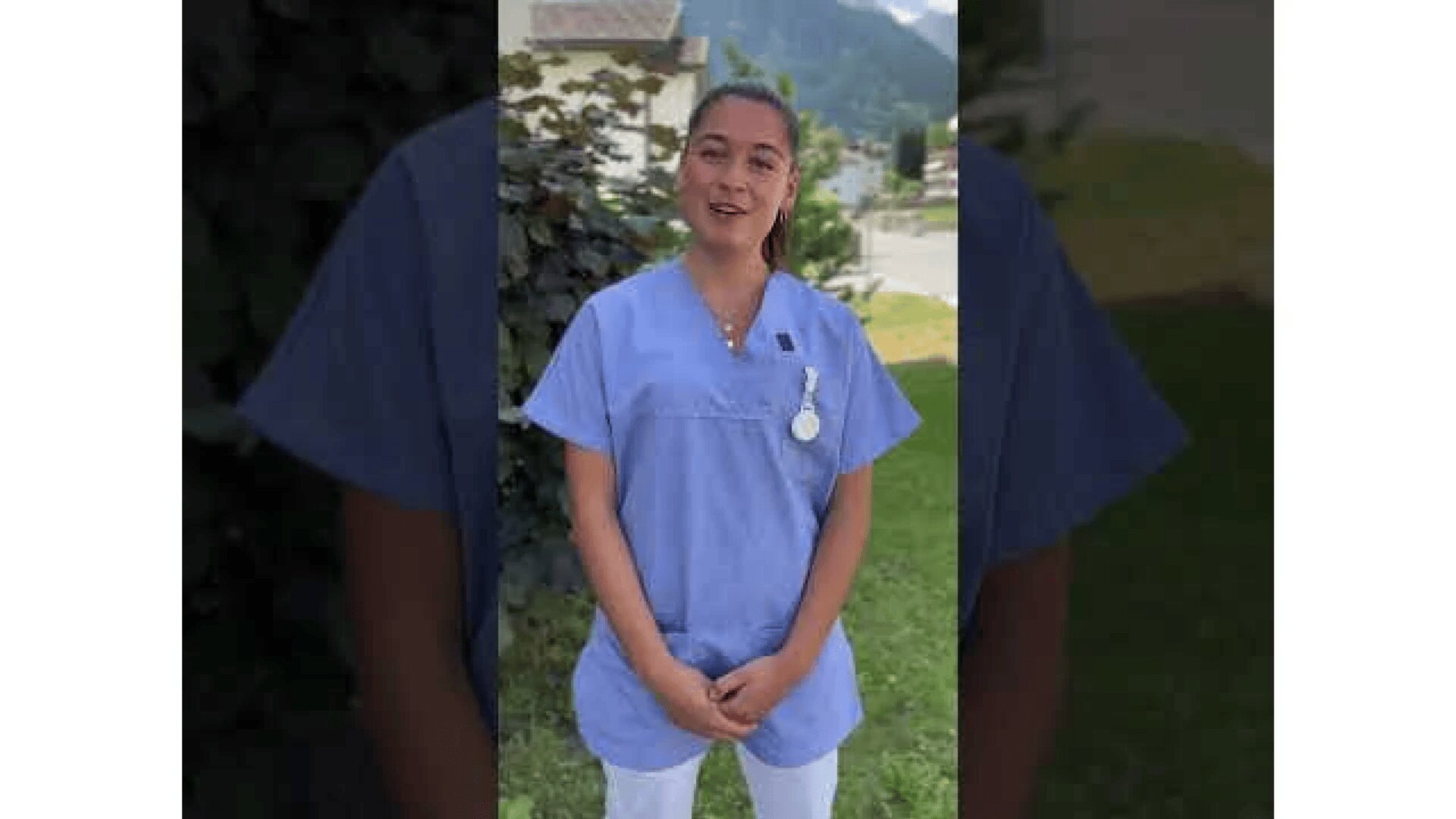 Eine Person, die einen hellblauen Arztkittel und ein Namensschild trägt, steht im Freien auf einer Wiese, mit Bäumen und einem Gebäude im Hintergrund.