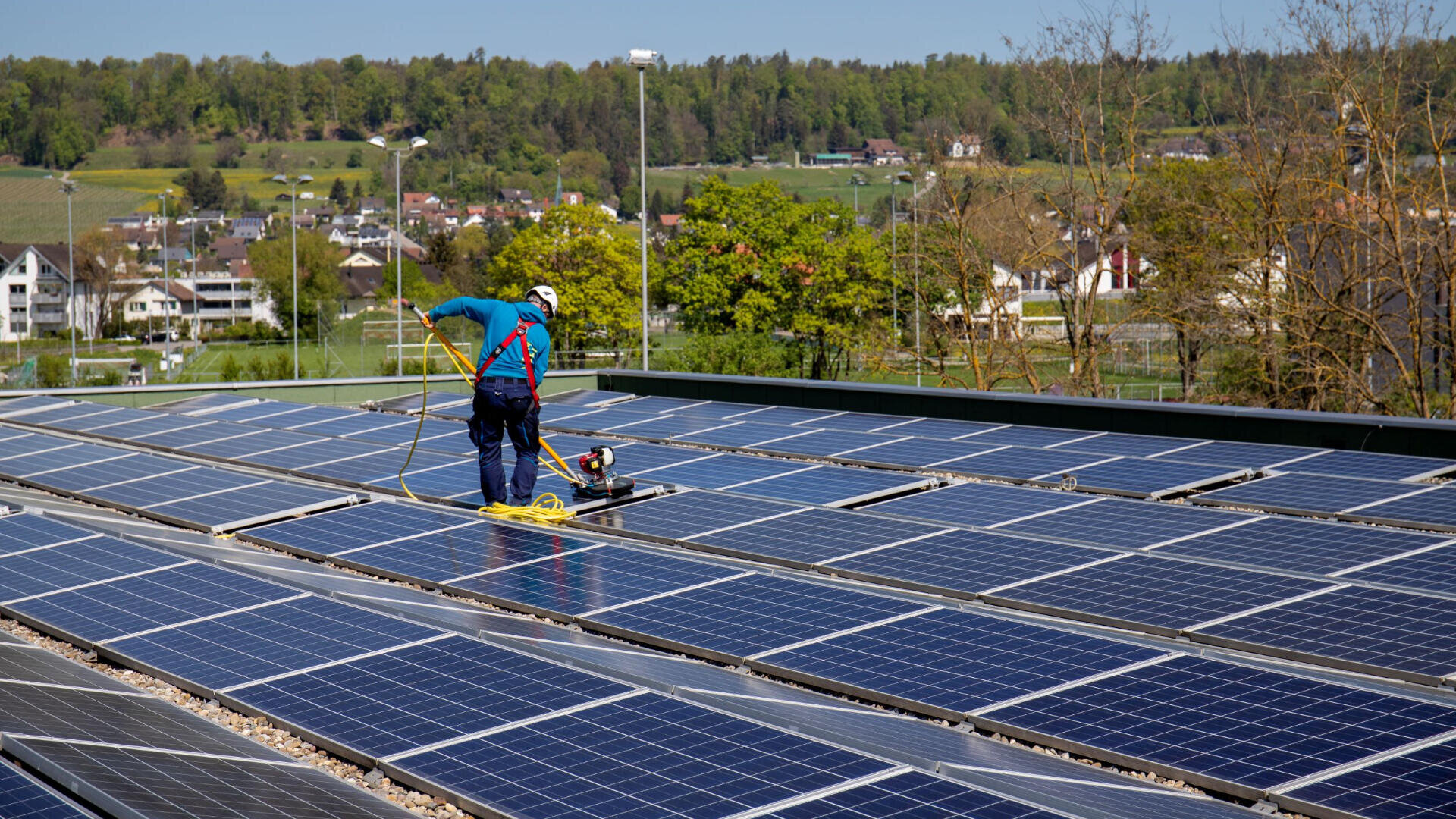 Eine Person in Schutzkleidung reinigt Solarmodule auf einem Dach mit Bürste und Schlauch. Im Hintergrund sind Grünflächen und Häuser unter einem leicht bewölkten Himmel zu sehen.