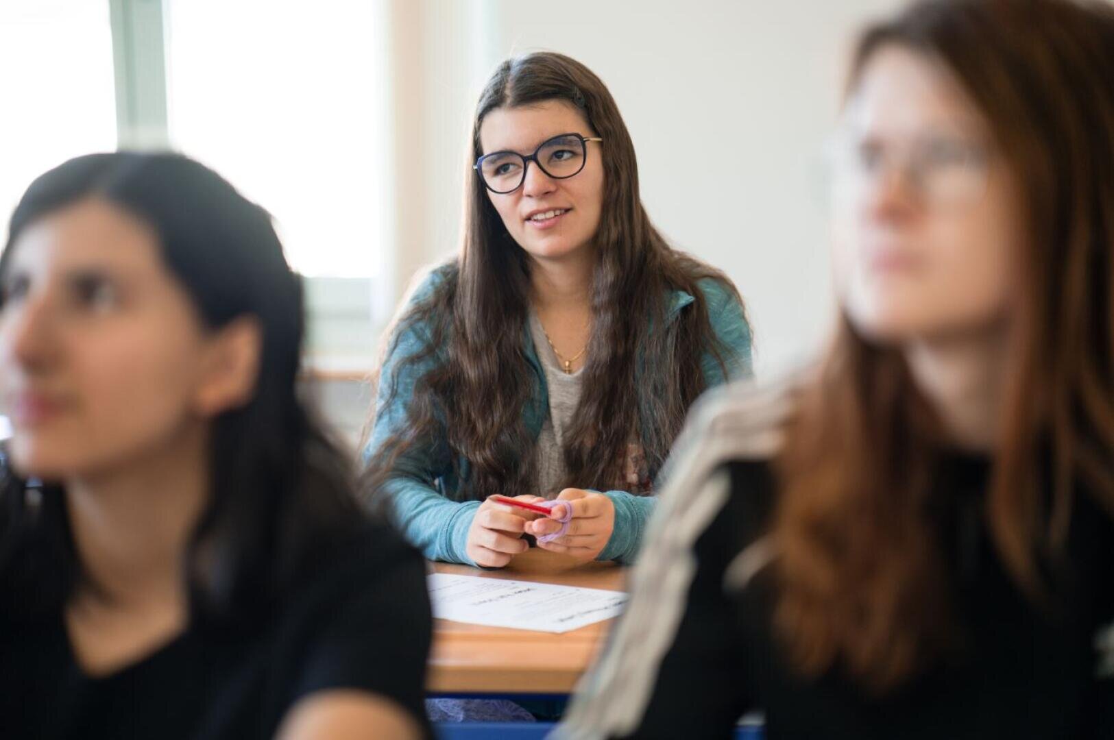 Eine junge Frau mit langen braunen Haaren und Brille sitzt an einem Schreibtisch in einem Klassenzimmer, hält einen roten Stift in der Hand und blickt nach vorne, während zwei andere Schüler im Vordergrund stehen.