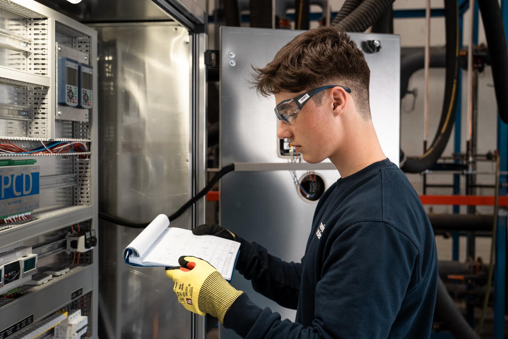 Ein junger Mann mit Schutzbrille und Handschuhen prüft ein Bedienfeld und macht sich Notizen auf einem Klemmbrett in einer industriellen oder technischen Anlage.