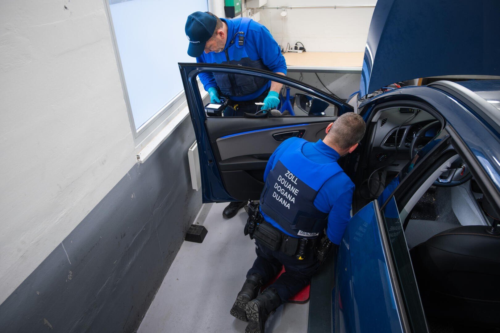 Zwei uniformierte Zollbeamte inspizieren den Innenraum und das Armaturenbrett eines blauen Autos in einer Garage. Ein Beamter kniet neben der offenen Fahrertür, während der andere die Papiere auf dem Autodach überprüft.