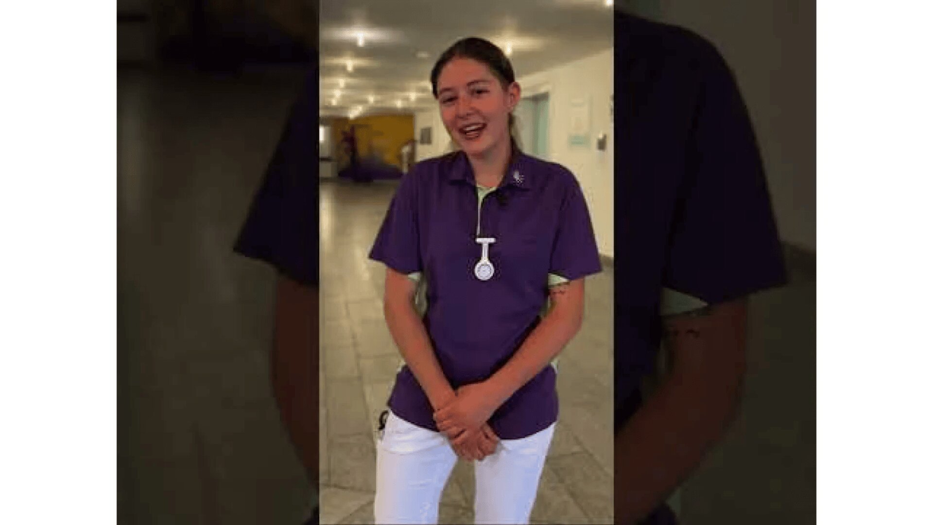 Eine Frau in einem lila Hemd und einer weißen Hose steht in einem Haus und lächelt in die Kamera. Sie hat ein Namensschild und eine Uhr an ihrem Hemd befestigt. Der Hintergrund ist ein Flur mit weicher Beleuchtung.