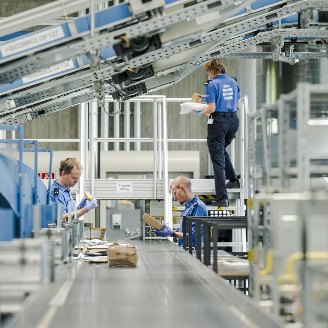 Drei Arbeiter in blauen Uniformen sortieren Pakete auf einem Förderbandsystem in einer Industrieanlage, wobei ein Arbeiter auf einer Plattform steht und zwei andere am Band darunter arbeiten.