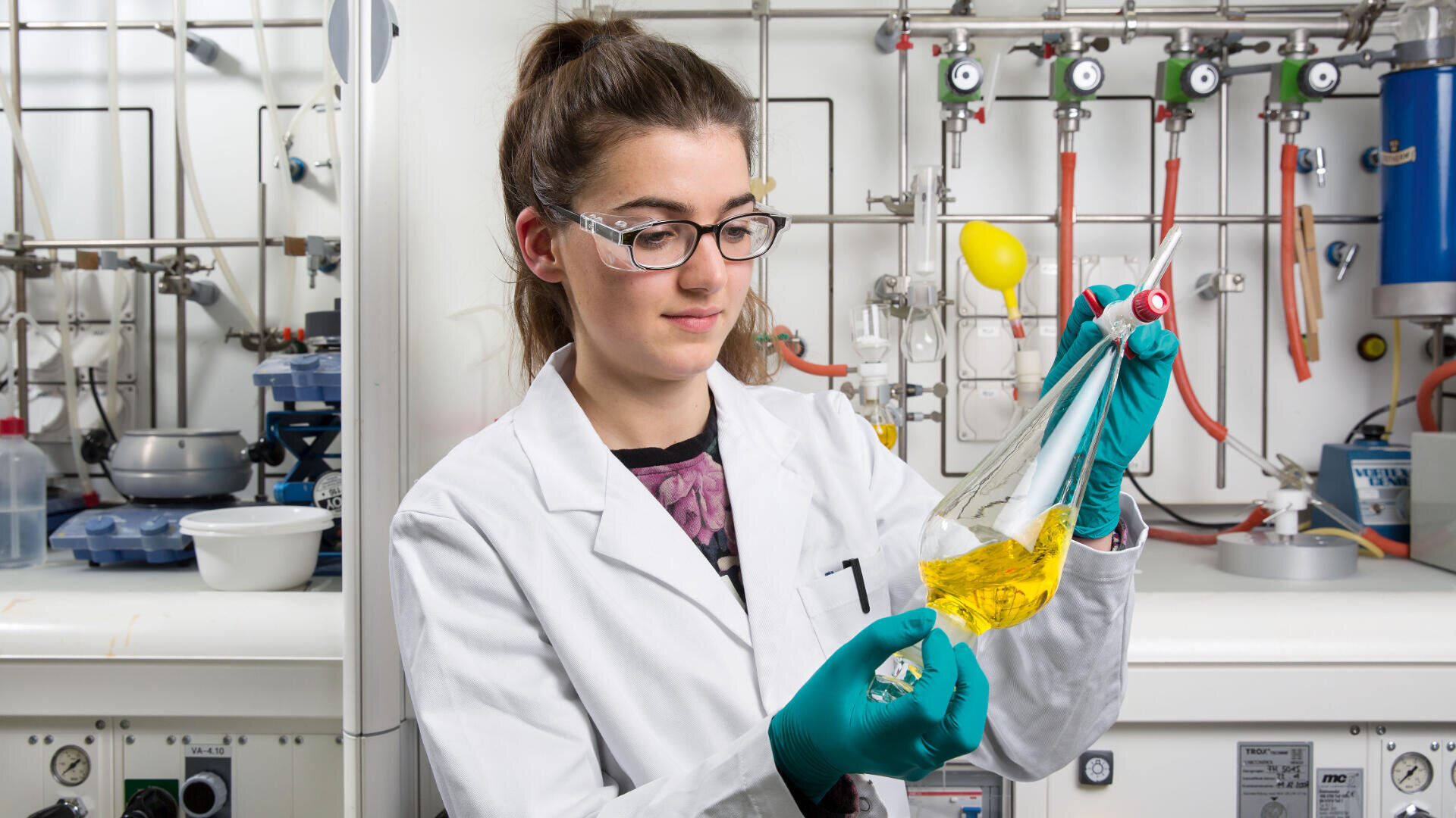 Eine Frau mit Laborkittel, Schutzbrille und blaugrünen Handschuhen untersucht einen Kolben mit einer gelben Flüssigkeit in einem Labor mit wissenschaftlichen Geräten und Rohren im Hintergrund.