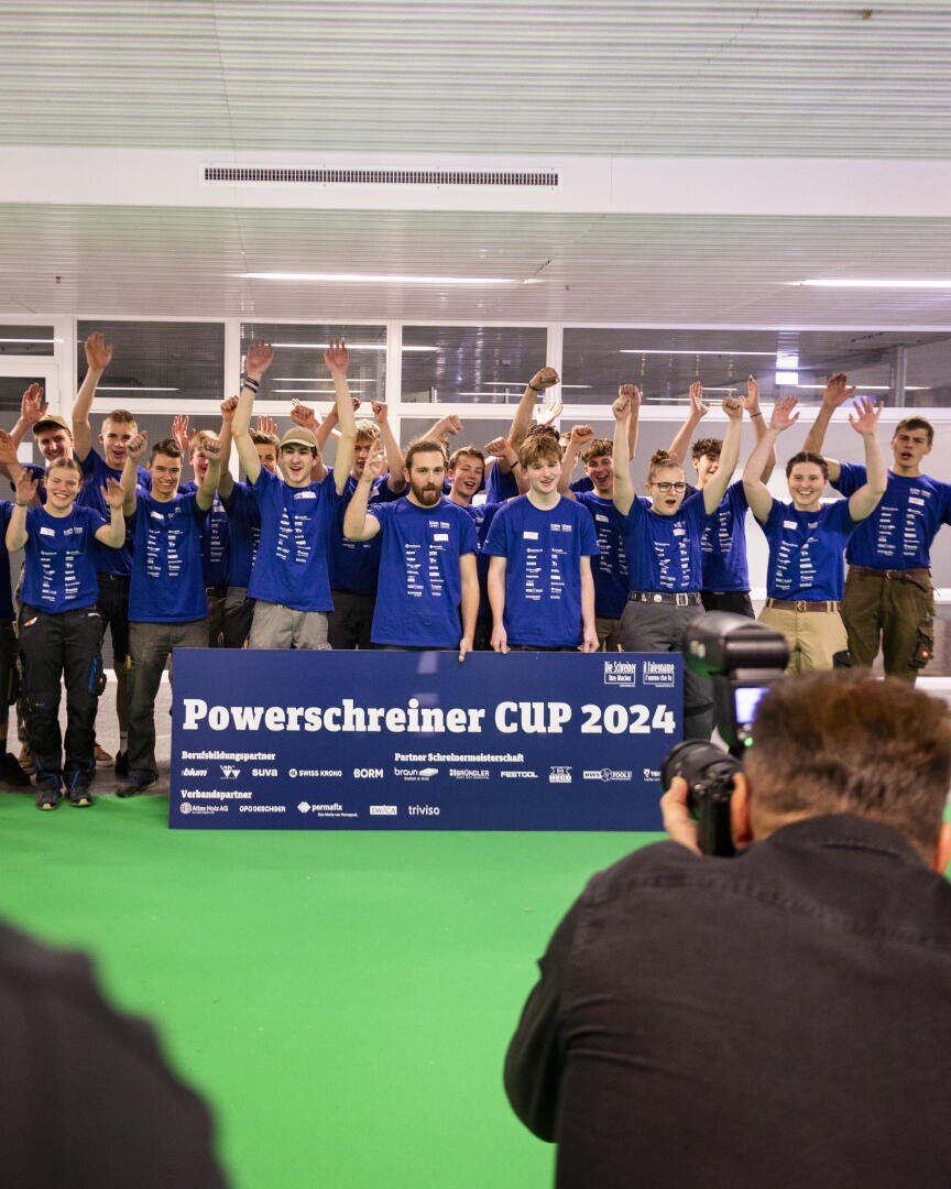 Eine Gruppe junger Leute in blauen Hemden jubelt mit erhobenen Armen hinter einem Schild mit der Aufschrift Powerschreiner CUP 2024, während Fotografen den Moment an der Front festhalten.