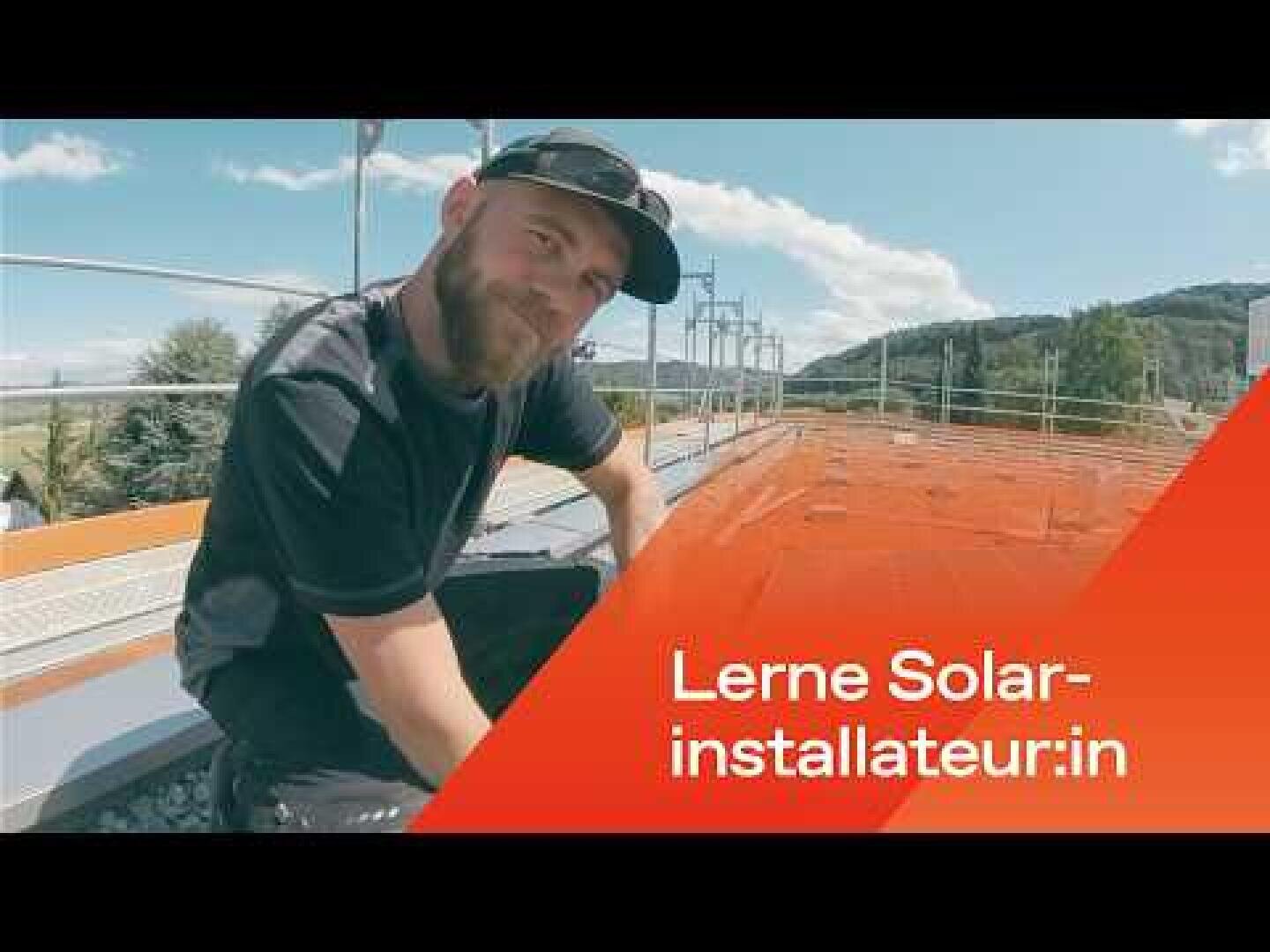 Eine Person mit Bart und Mütze sitzt auf einem Dach mit Solarmodulen und lächelt in die Kamera. Der Text lautet: „Lerne Solarinstallateur:in“ auf orangefarbenem Hintergrund. Im Hintergrund sind Bäume und Hügel zu sehen.
