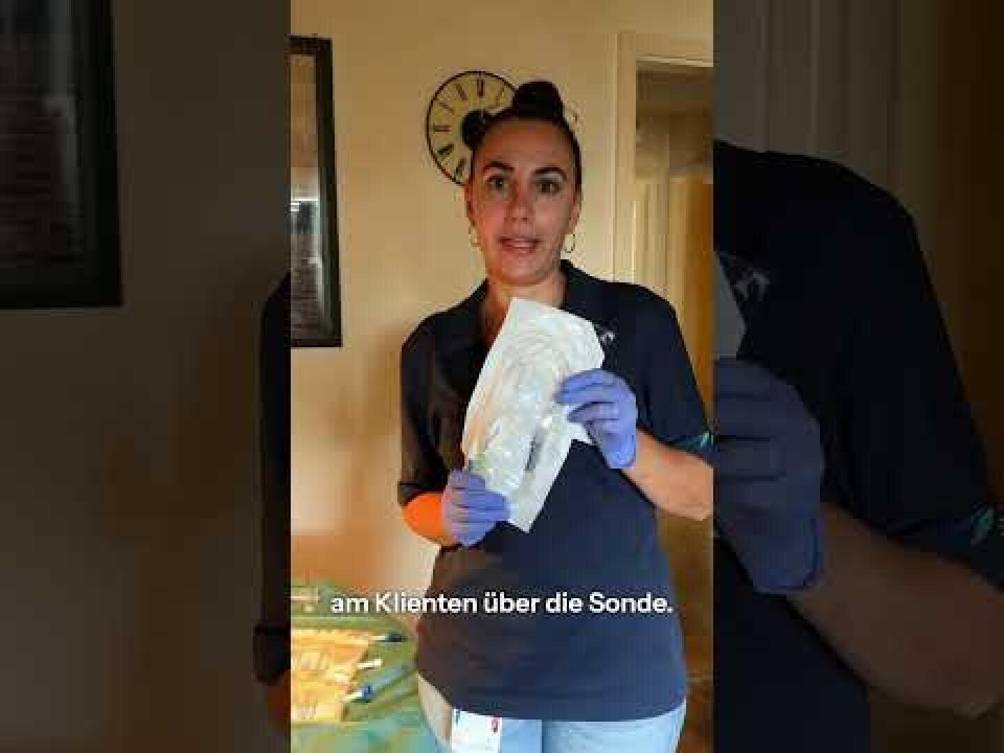 Eine Frau mit Handschuhen hält ein medizinisches Gerät in der Hand und steht in einem Raum, hinter ihr an der Wand eine Uhr. Die deutschen Untertitel am unteren Bildrand lauten "Am Klienten über die Sonde".