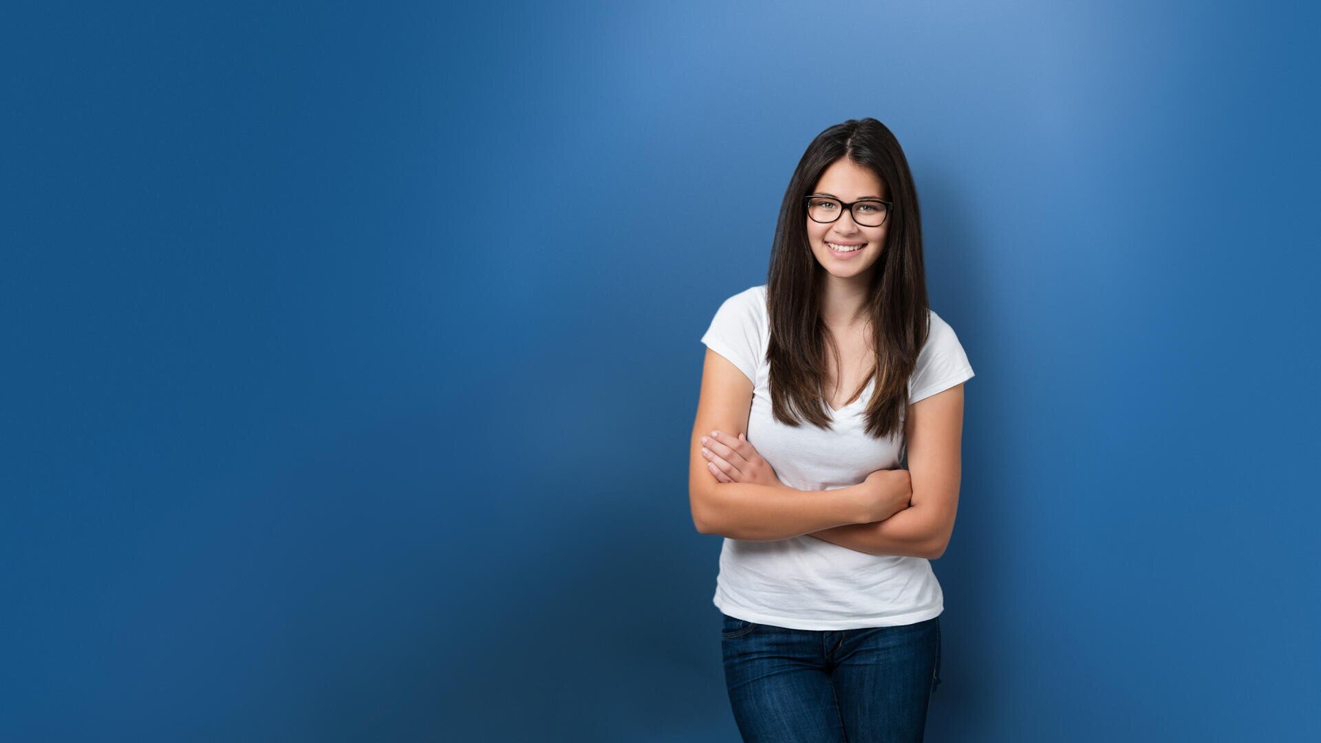 Eine junge Frau mit langen dunklen Haaren, Brille, weißem T-Shirt und Bluejeans steht lächelnd mit verschränkten Armen vor einem schlichten blauen Hintergrund.