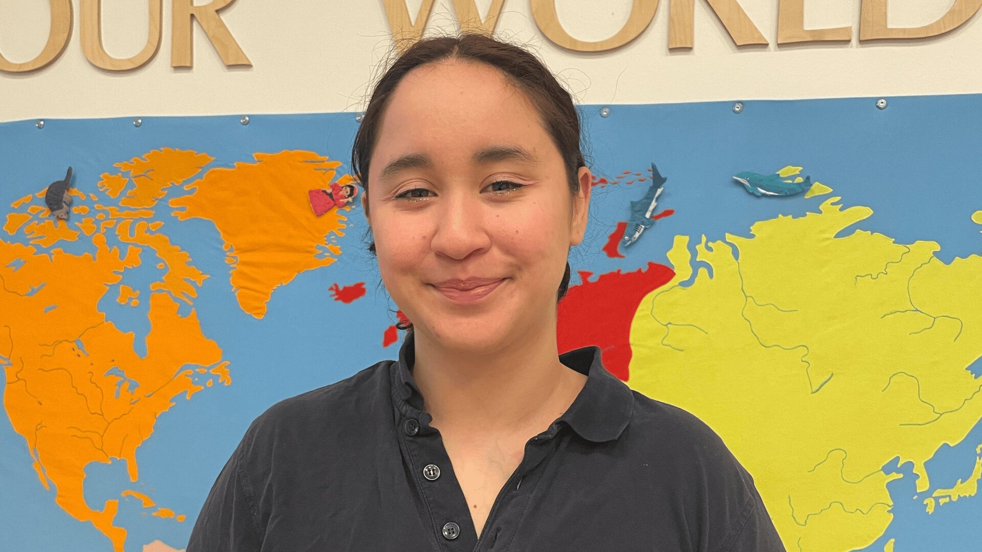 Eine Person mit zurückgebundenem dunklem Haar und schwarzem Kragenhemd lächelt vor einer bunten Weltkarte an der Wand. Die Worte OUR WORLD sind teilweise über ihnen sichtbar.