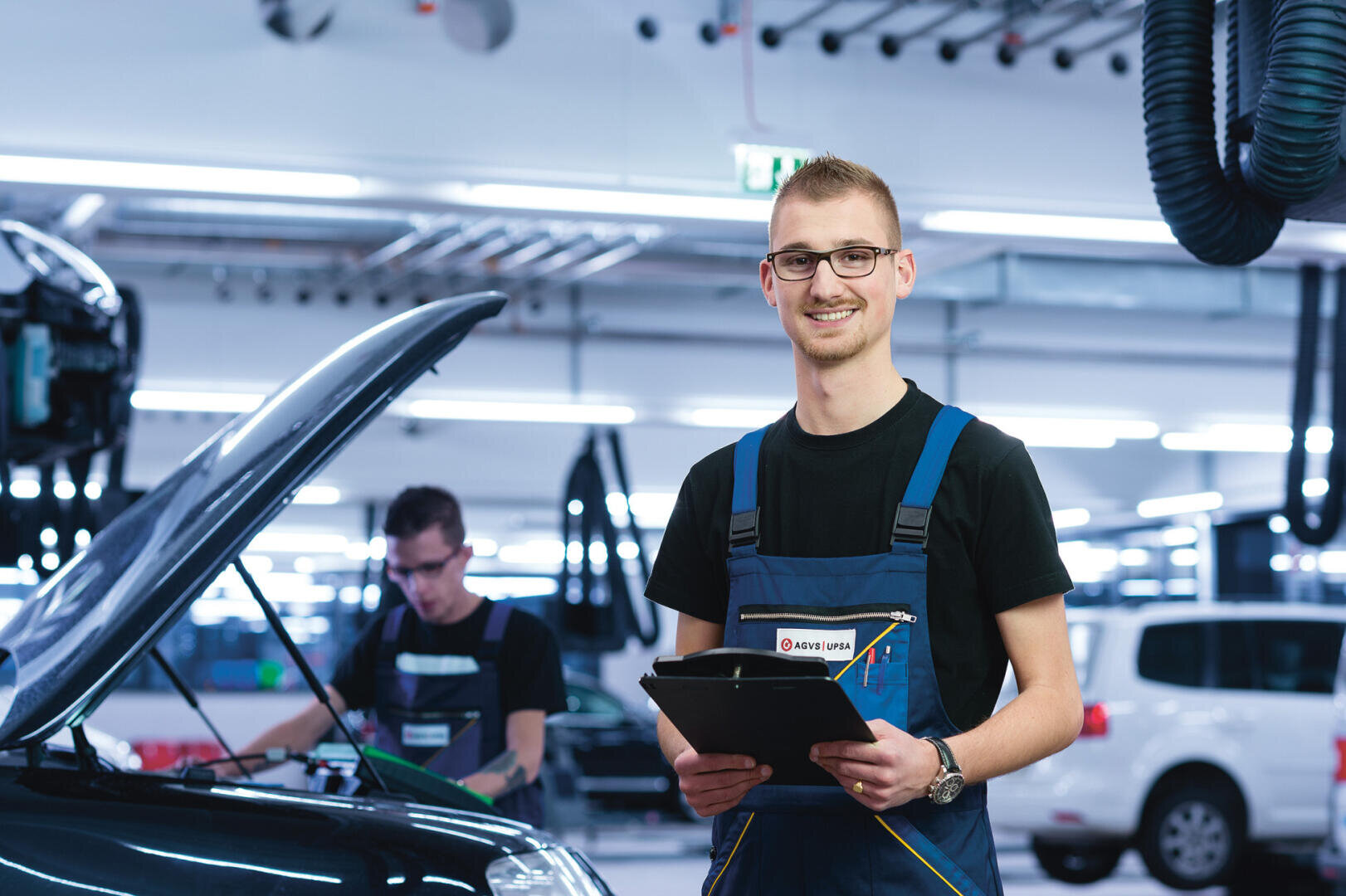 Ein lächelnder Kfz-Mechaniker in blauer Arbeitskleidung hält in einer Werkstatt ein Klemmbrett in der Hand, während im Hintergrund ein anderer Techniker an einem Auto arbeitet.
