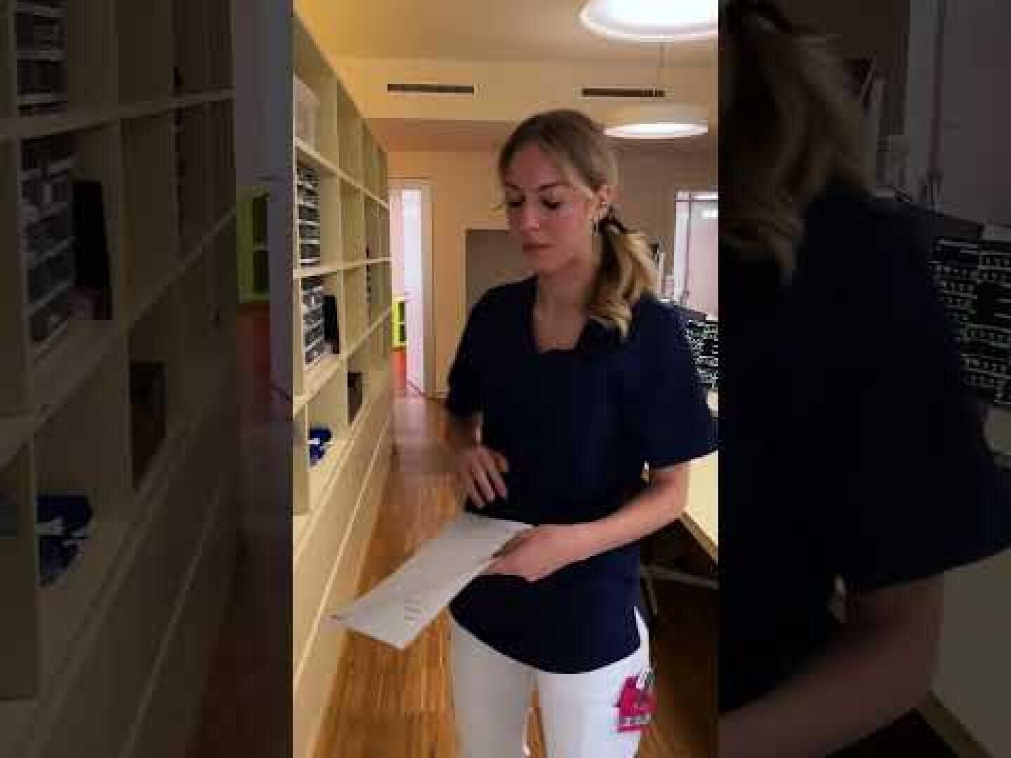 Eine Frau in medizinischer Kleidung steht in einem mit Regalen gesäumten Flur und hält ein Stück Papier in der Hand. Die Umgebung scheint eine medizinische oder Büroumgebung zu sein, mit Holzfußböden und Oberlichtern.