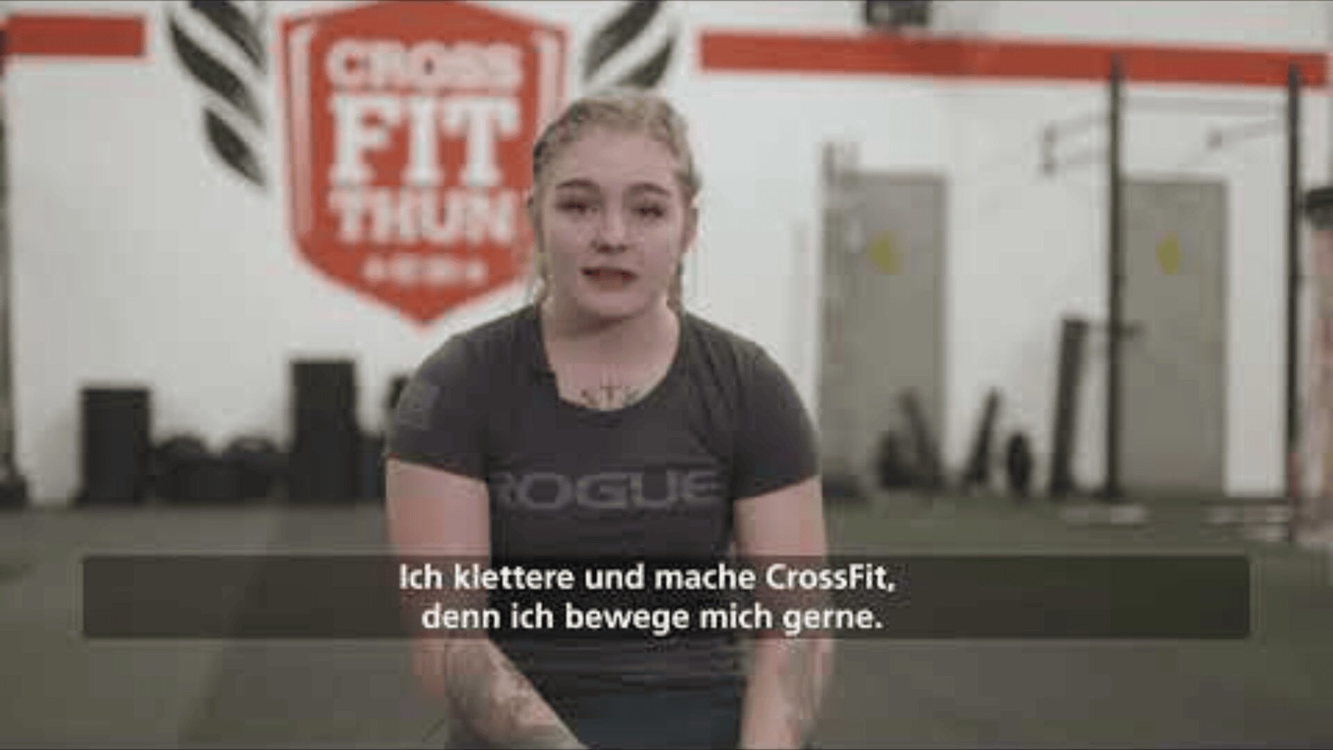 Eine Frau mit blonden Haaren im Pferdeschwanz und Tattoos sitzt in einem Fitnessstudio und spricht in die Kamera. Hinter ihr ist ein CrossFit Thun-Logo zu sehen. Die deutschen Untertitel lauten: Ich klettere und mache CrossFit, denn ich bewege mich gerne.