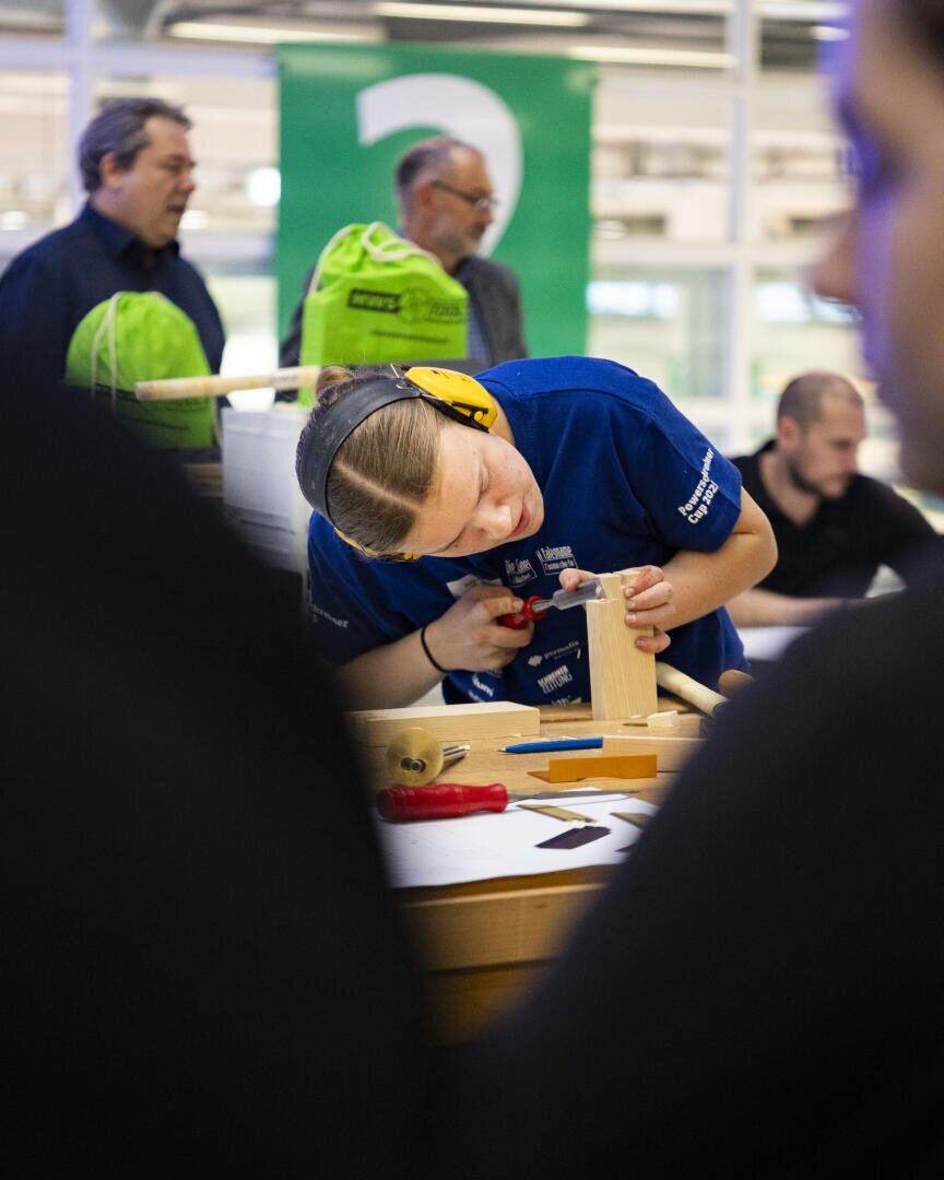 Eine Person in einem blauen Hemd und mit Gehörschutz arbeitet mit einem Werkzeug an einem Holzprojekt an einer Werkbank, während im Hintergrund andere Personen und grüne Taschen zu sehen sind.