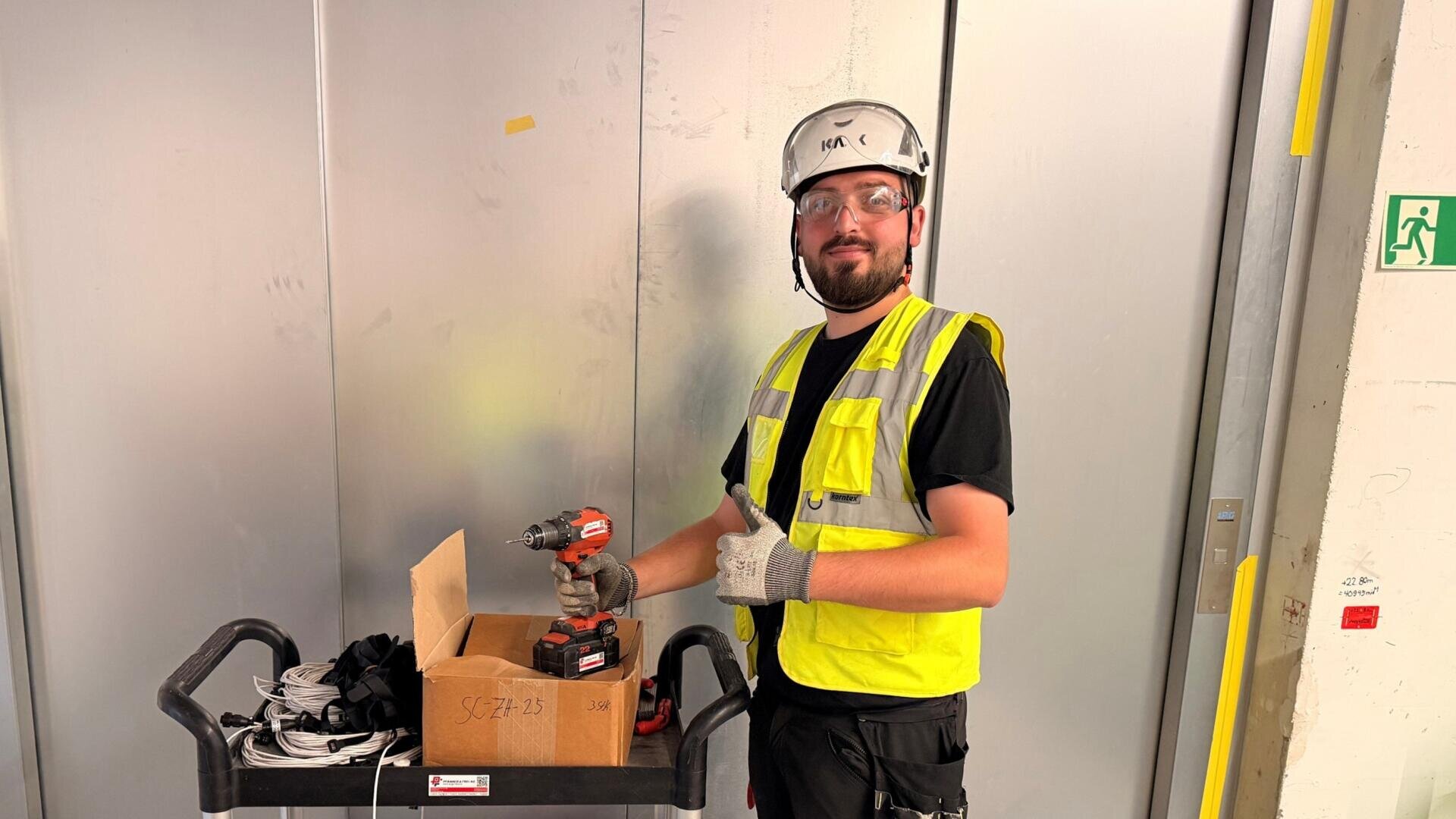 Ein Bauarbeiter, der einen Schutzhelm, eine Schutzbrille und eine gelbe Weste trägt, steht neben einem Wagen mit Werkzeugen, hält eine Bohrmaschine in der Hand und streckt vor einer Metallwand den Daumen nach oben.