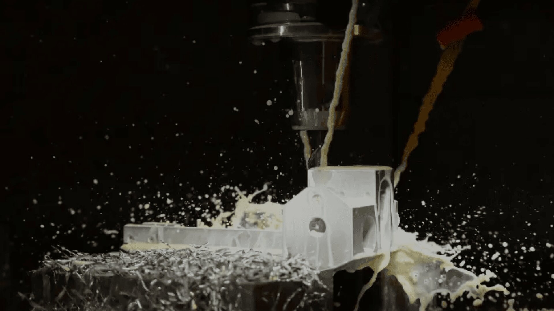 Eine CNC-Maschine bohrt in einen Metallblock, wobei Kühlflüssigkeit versprüht wird und Metallspäne vor einem dunklen Hintergrund auf der Arbeitsfläche verstreut sind.