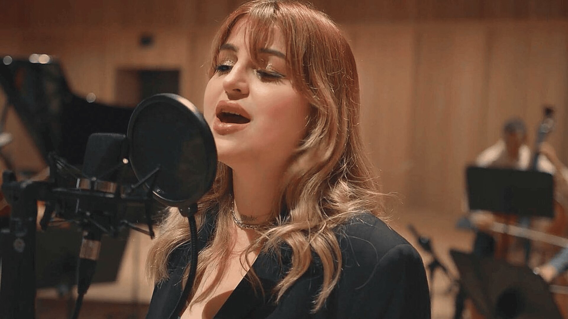 Eine Frau mit hellbraunem Haar singt in ein Studiomikrofon, während im verschwommenen Hintergrund eines holzgetäfelten Aufnahmestudios Musiker Streichinstrumente spielen.