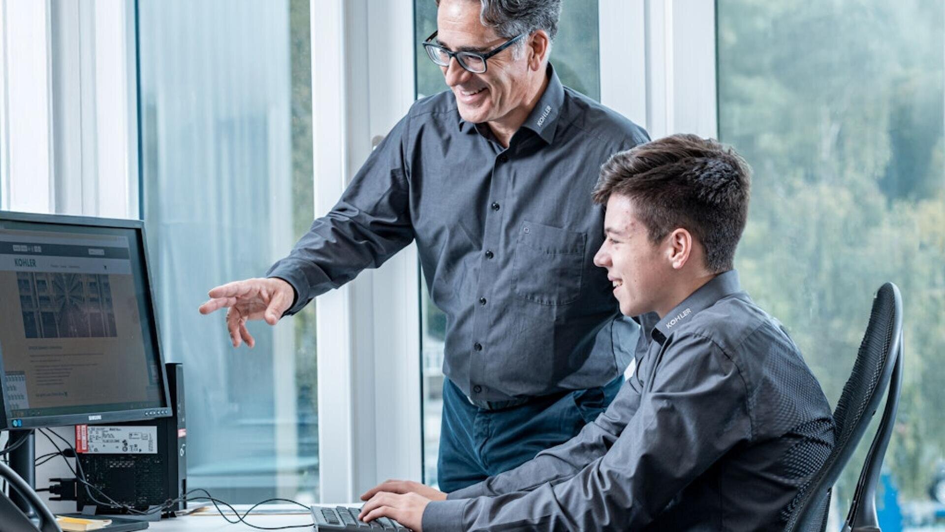 Zwei Personen in grauen Hemden arbeiten an einem Computer in einem hellen Büro. Die eine sitzt, tippt und lächelt, während die andere neben ihr steht, auf den Bildschirm zeigt und ebenfalls lächelt. Im Hintergrund sind große Fenster zu sehen.