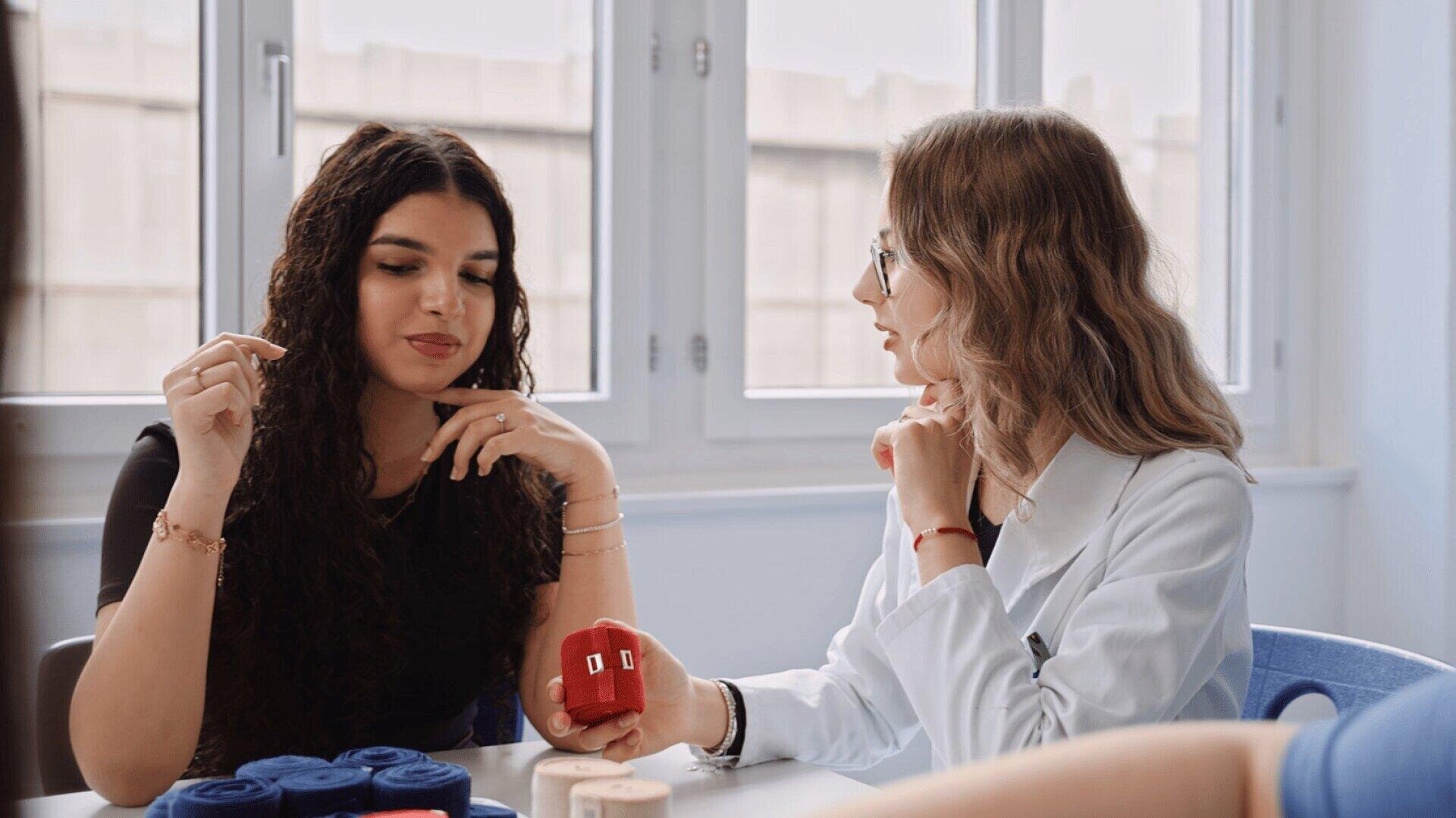 Zwei Frauen sitzen an einem Tisch in der Nähe großer Fenster. Die eine, in einem weißen Kittel, hält ein kleines rotes Modell in der Hand und gestikuliert, während die andere aufmerksam zuhört. Vor ihnen auf dem Tisch liegen Garnrollen.