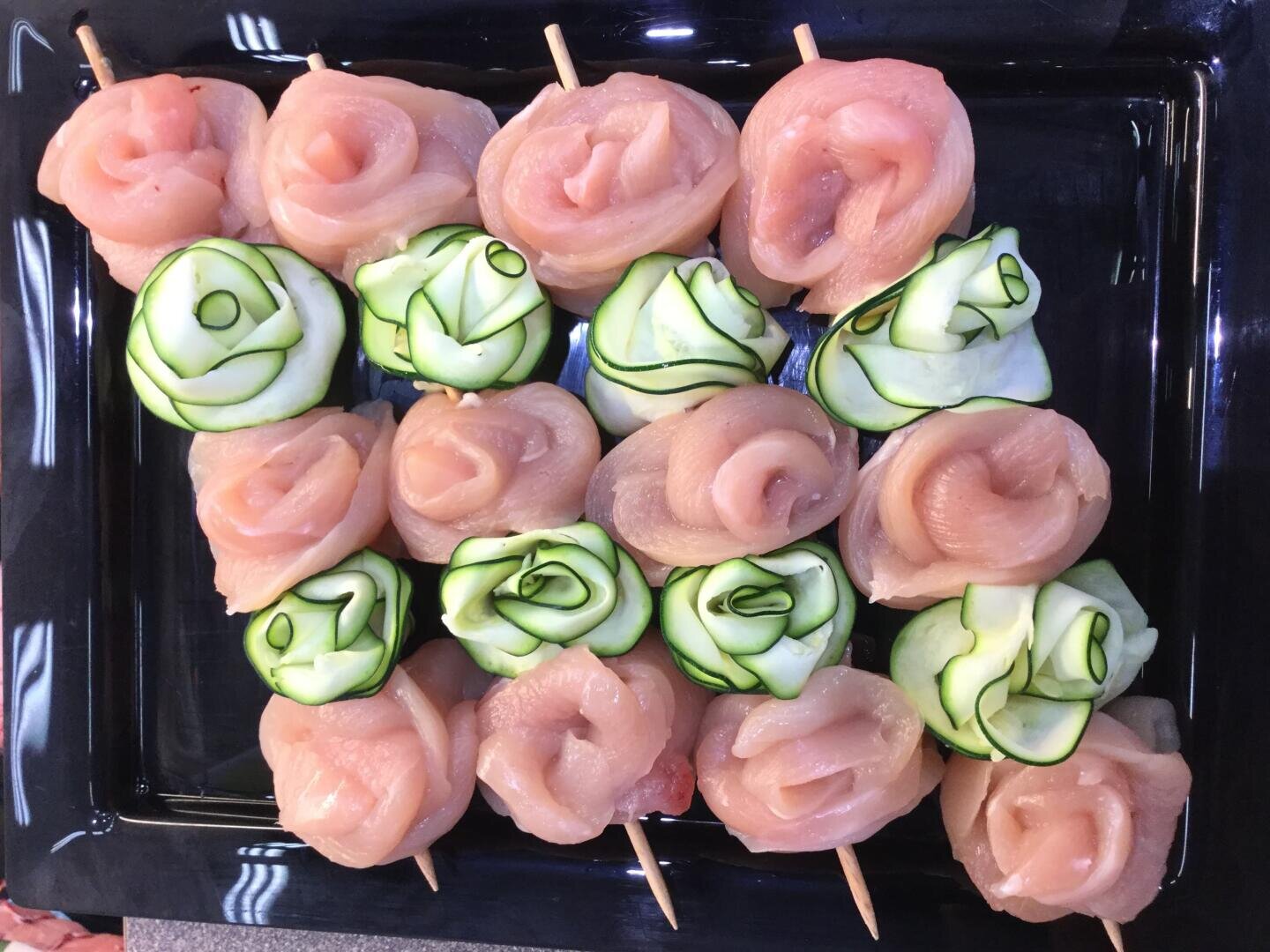 Rohe Hähnchenteile und dünne Zucchinischeiben werden auf einem schwarzen, rechteckigen Tablett aufgespießt und rosenförmig präsentiert.