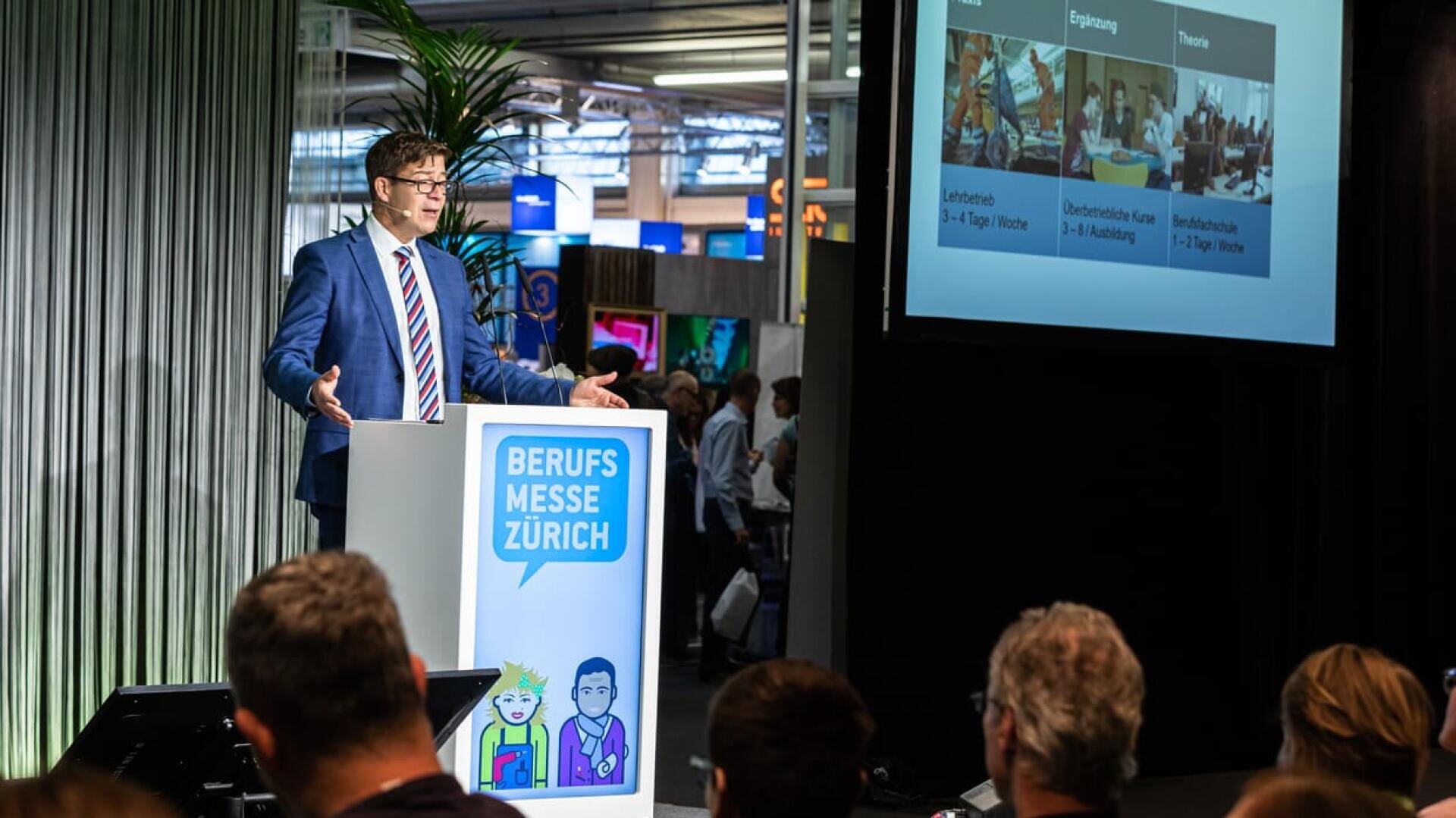 Ein Mann in einem blauen Anzug spricht an einem Podium mit der Aufschrift Berufsmesse Zürich vor einem Publikum, hinter ihm ein grosser Bildschirm mit Informationen und Bildern.