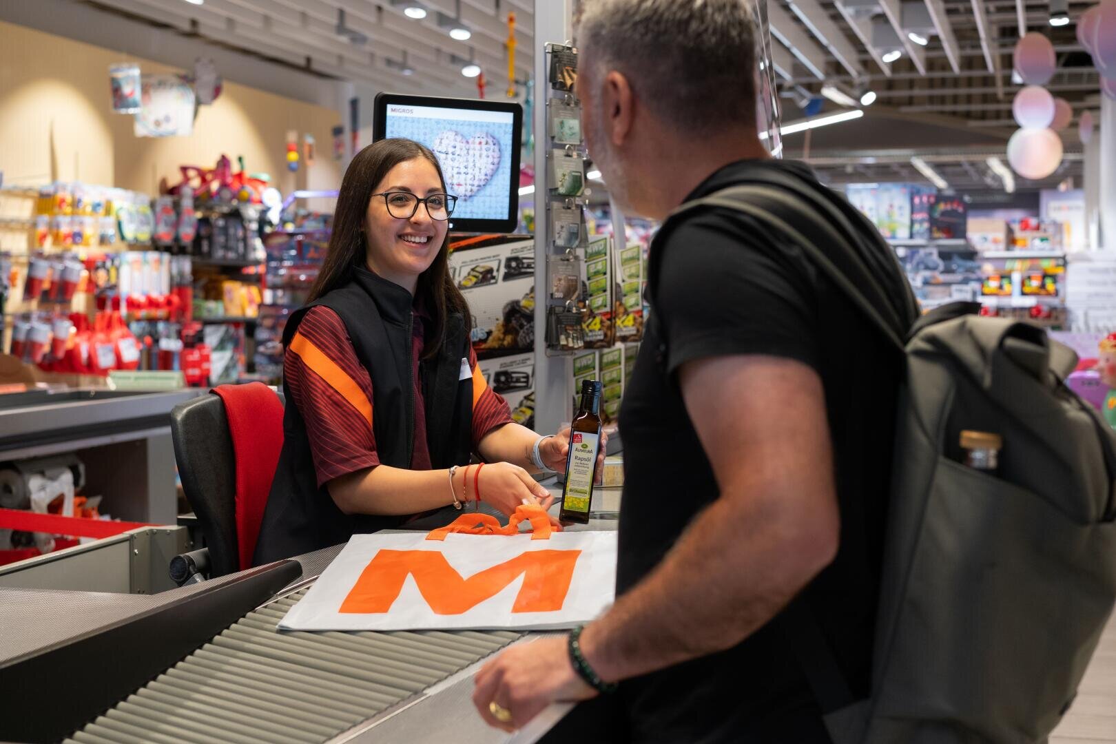 Eine lächelnde Kassiererin überreicht einem Kunden an der Supermarktkasse eine Flasche. Der Kunde hat einen Rucksack und eine wiederverwendbare Tasche mit einem großen orangefarbenen M auf dem Förderband. Im Hintergrund sind Regale mit verschiedenen Produkten zu sehen.