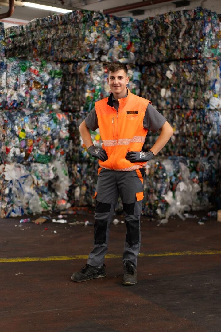 Ein Arbeiter in einer orangefarbenen Sicherheitsweste und mit Handschuhen steht in einer Recyclinganlage vor großen, zusammengepressten Stapeln aus recyceltem Kunststoff.