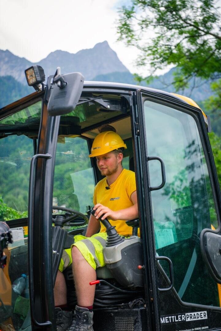 Ein Bauarbeiter, der einen gelben Schutzhelm und reflektierende Shorts trägt, bedient schwere Maschinen in einer Kabine, während im Hintergrund Berge und Bäume zu sehen sind.
