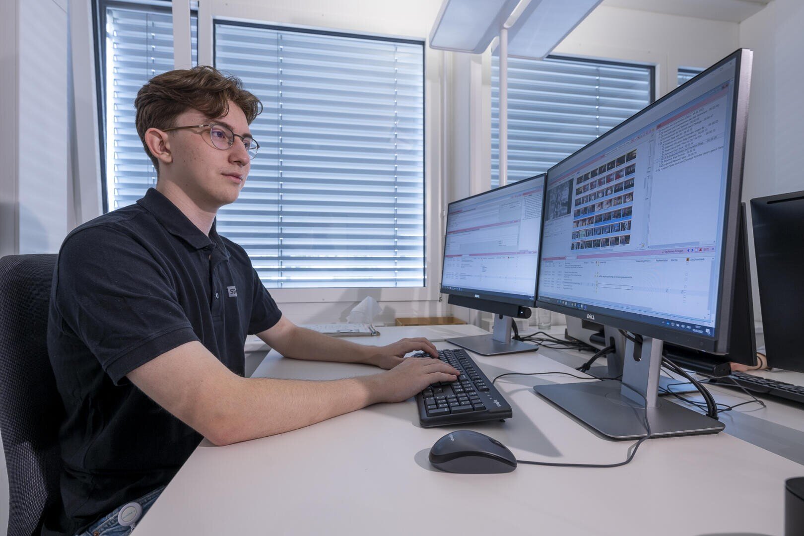 Ein junger Mann mit Brille sitzt an einem Schreibtisch und arbeitet an einem Computer mit zwei großen Monitoren, auf denen Software mit Texten und Bildern angezeigt wird. Jalousien verdecken die Fenster hinter ihm, und das Büro wirkt hell und modern.