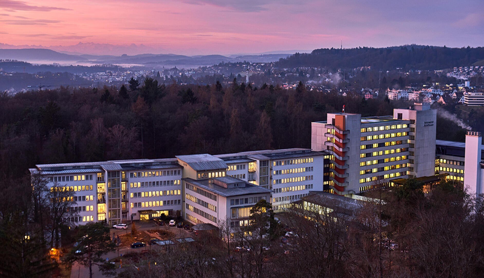 Ein großer, moderner Krankenhauskomplex ist in der Abenddämmerung hell erleuchtet, umgeben von Bäumen und mit einer Stadtlandschaft und Bergen im Hintergrund unter einem lila-rosa Himmel.
