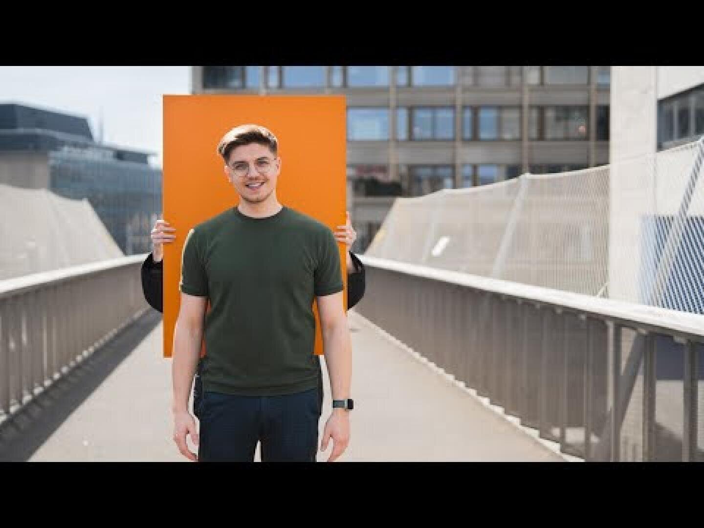 Ein junger Mann in einem grünen T-Shirt steht lächelnd auf einer Fußgängerbrücke, während eine andere Person hinter ihm ein großes, orangefarbenes, rechteckiges Schild hochhält, das teilweise nicht zu sehen ist.