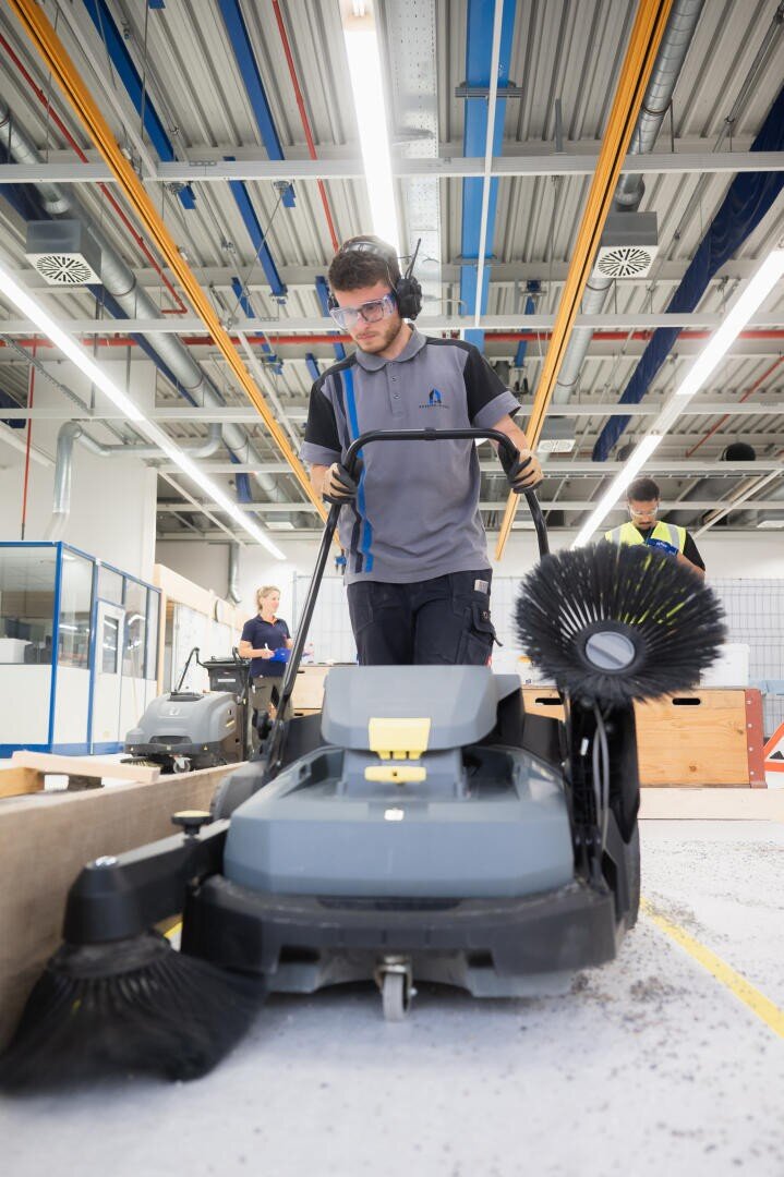 Eine Person, die eine Schutzbrille und Arbeitskleidung trägt, bedient eine große Bodenreinigungsmaschine in einer Industrieumgebung, während im Hintergrund ein weiterer Arbeiter und Reinigungsgeräte zu sehen sind.