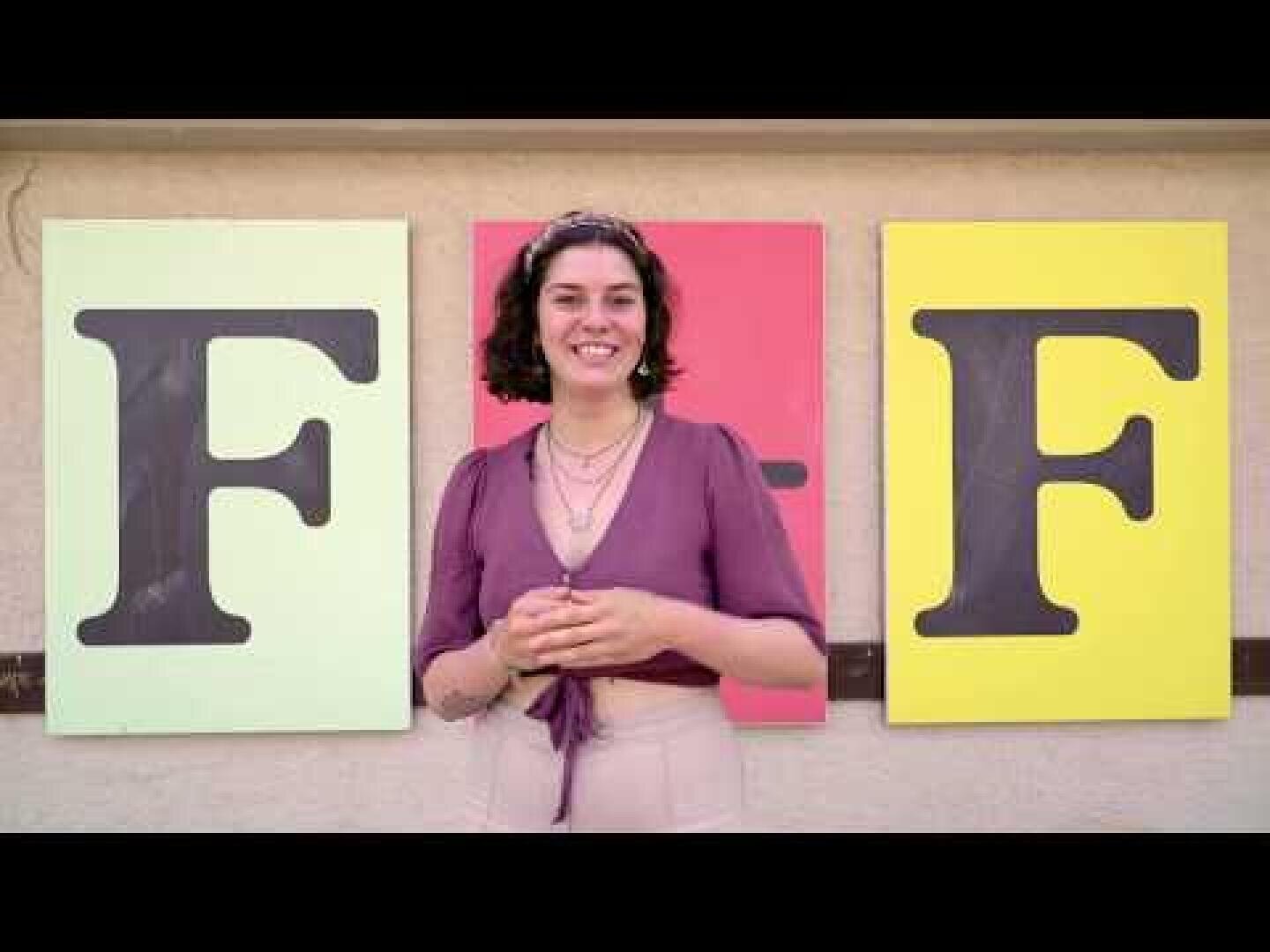 Eine lächelnde Person mit kurzen dunklen Haaren steht vor drei großen Schildern. Die Schilder zeigen die Buchstaben F, - und F auf grünem, rotem und gelbem Hintergrund. Die Person trägt ein lila Oberteil und eine beigefarbene Hose.