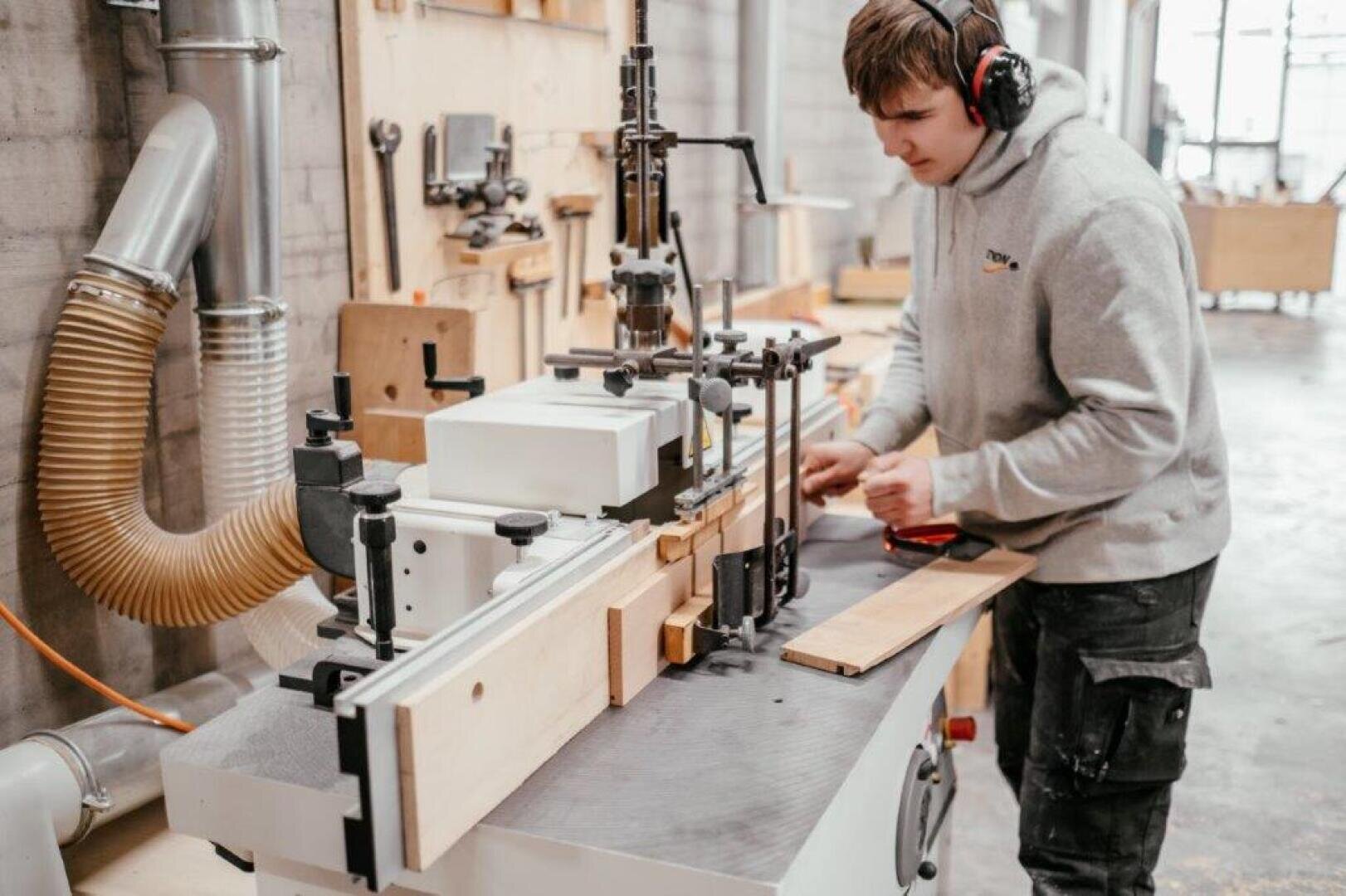 Eine Person mit Gehörschutz bedient eine Holzbearbeitungsmaschine in einer Werkstatt und führt ein Holzbrett durch die Anlage. Im Hintergrund sind verschiedene Werkzeuge, Schläuche und Materialien zu sehen.