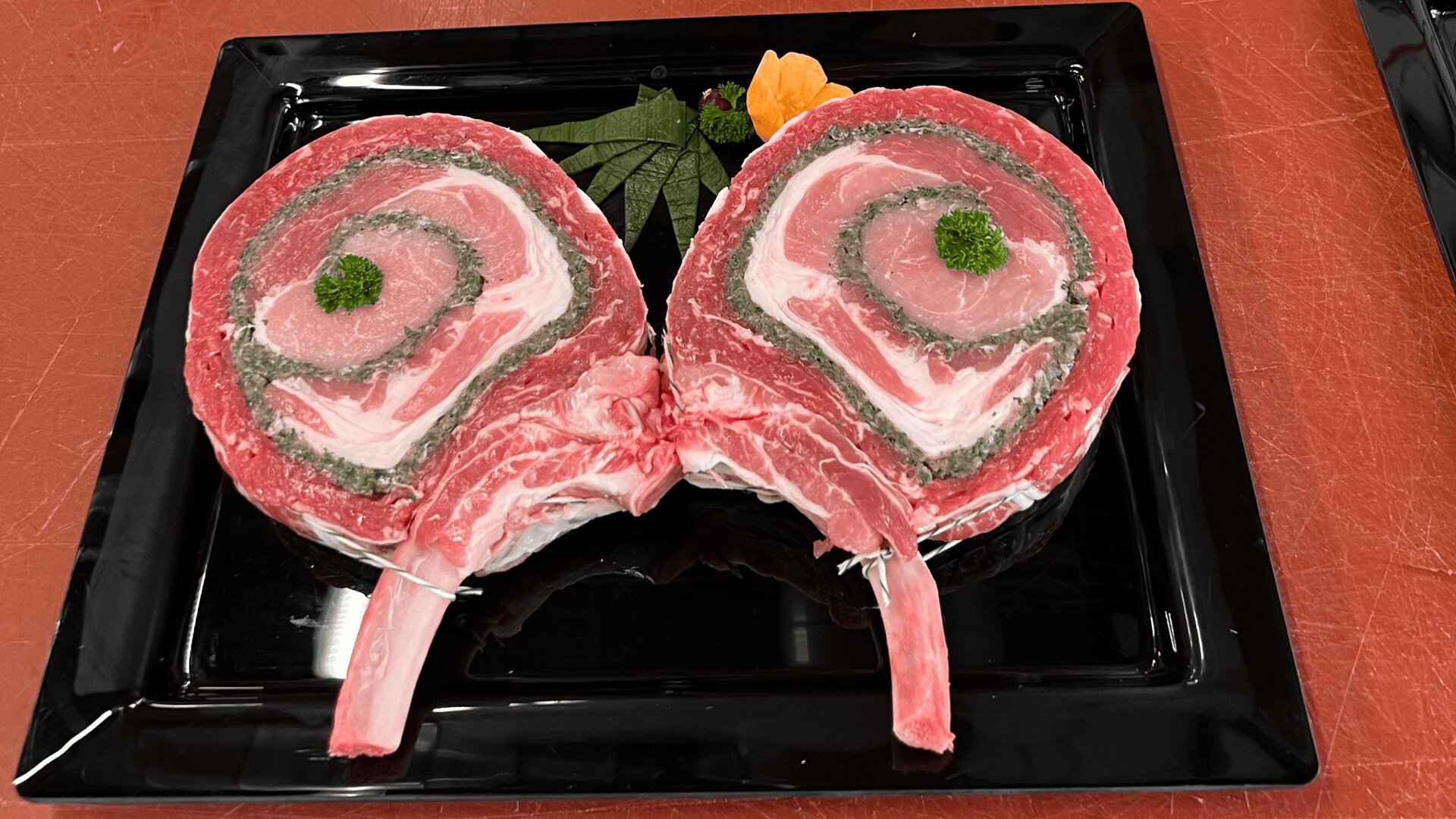 Zwei gerollte, gefüllte, rohe Fleischstücke mit Knochen werden auf einem schwarzen Tablett in Form von Schmetterlingsflügeln angerichtet und mit Petersilie und einer kleinen Orangenblüte garniert.