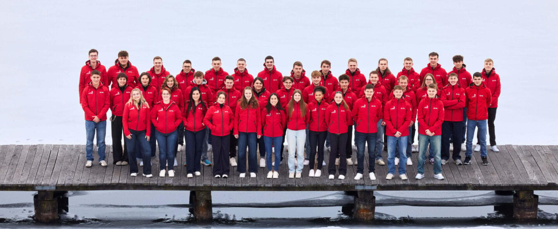 Eine Gruppe von etwa 50 Personen in einheitlichen roten Jacken steht auf einem Holzsteg über ruhigem Wasser und posiert für ein Gruppenfoto. Der Hintergrund ist neblig und grau, was auf einen kalten Tag hindeutet.