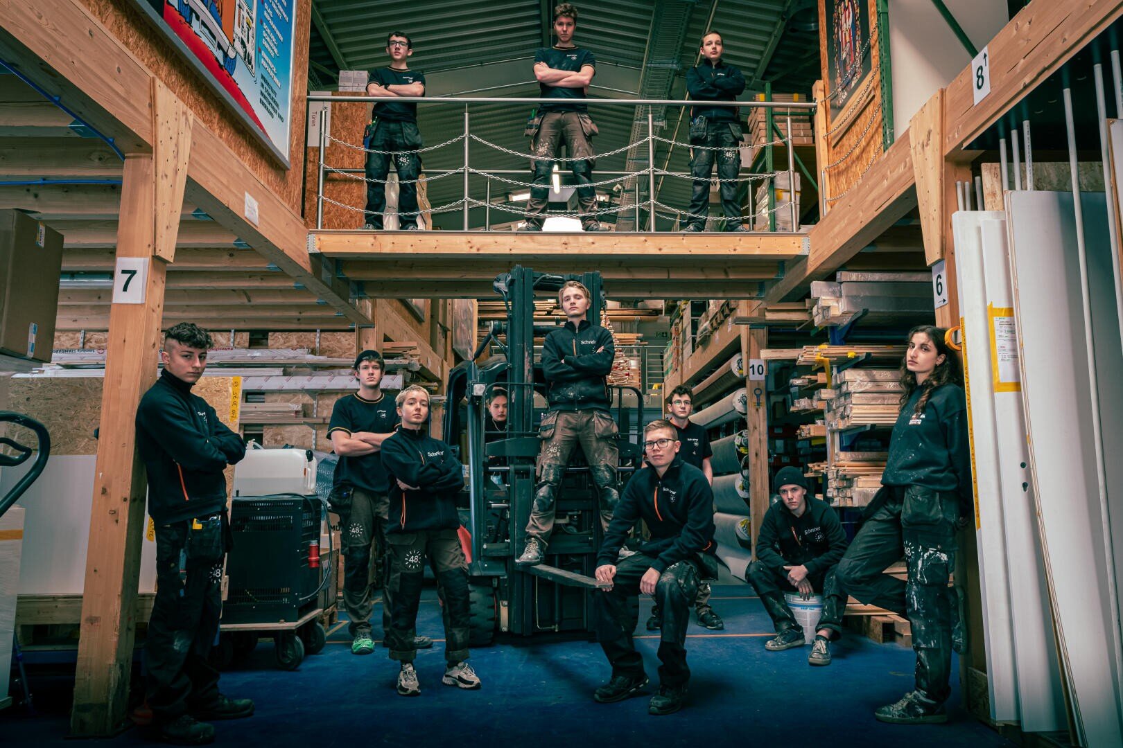 Eine Gruppe von Arbeitern in dunklen Uniformen posiert in einem Lagerhaus, einige stehen im Erdgeschoss, andere auf einem oberen Zwischengeschoss. Um sie herum stehen Regale mit Materialien und Geräten, die eine industrielle Atmosphäre schaffen.