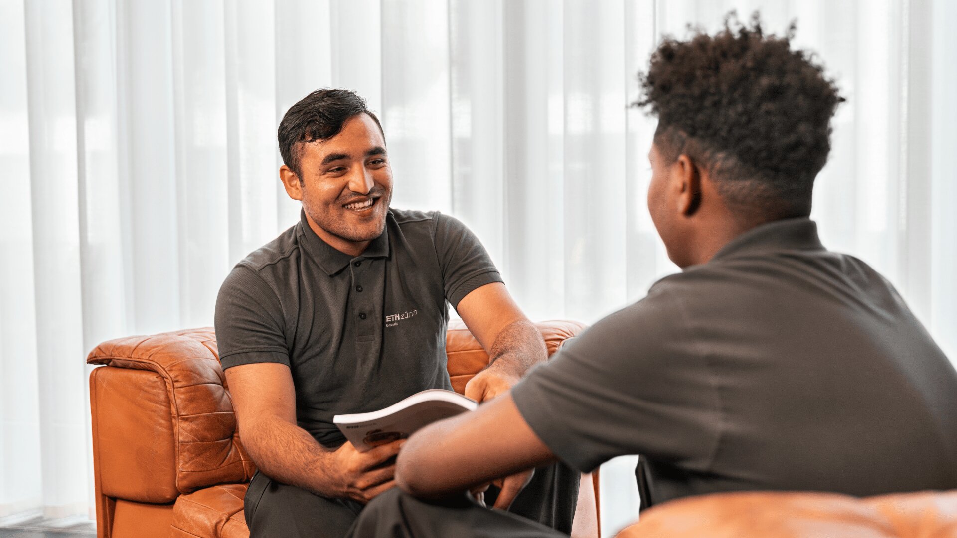 Zwei Männer in grauen Poloshirts sitzen sich auf braunen Lederstühlen gegenüber. Einer lächelt und hält eine Broschüre in der Hand, während sie sich in einem hellen Raum mit weißen Vorhängen im Hintergrund unterhalten.