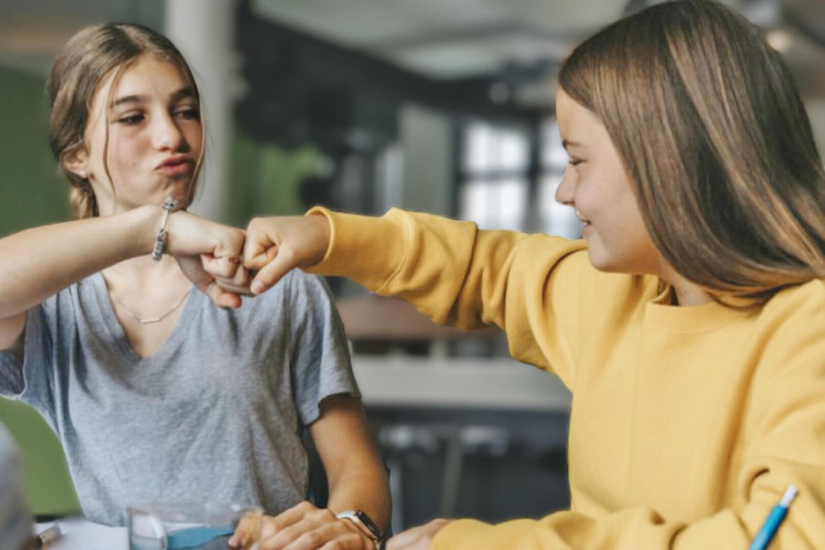 Zwei Mädchen sitzen drinnen an einem Tisch, lächeln und geben sich gegenseitig einen Faustschlag. Die eine trägt ein graues Hemd und die andere ein gelbes Sweatshirt. Der Hintergrund ist sanft verschwommen.