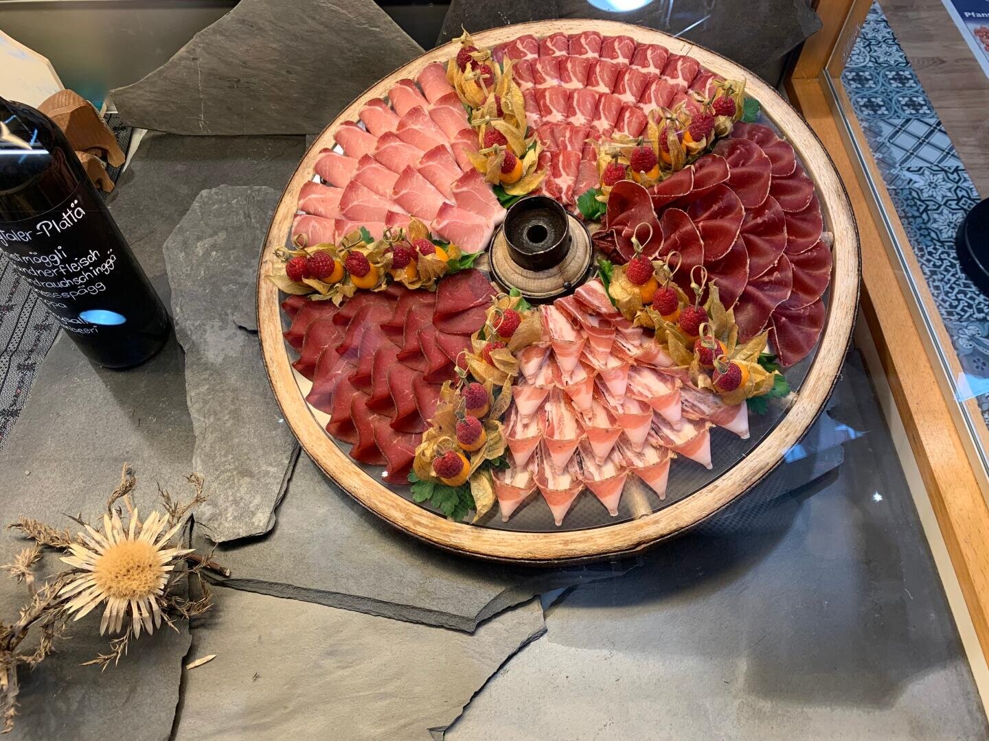 Eine runde Holzplatte mit verschiedenen dünn geschnittenen Wurstsorten, garniert mit gelben Blüten und Himbeeren, steht auf einer Schieferplatte neben einer Flasche Wein und einer getrockneten Distel.