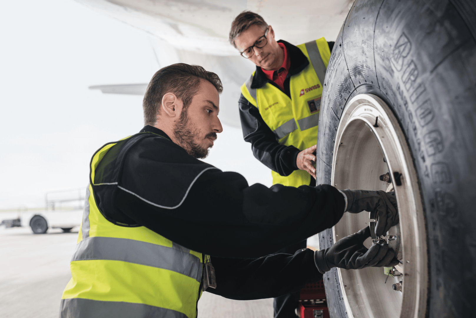 Zwei Mitarbeiter des Bodenpersonals eines Flughafens in gelben Sicherheitswesten inspizieren und arbeiten an einem Flugzeugreifen, wobei sie sich auf die Radbolzen konzentrieren. Ein Mann hockt in der Hocke, während der andere beobachtet, beide unter dem Flugzeugflügel.