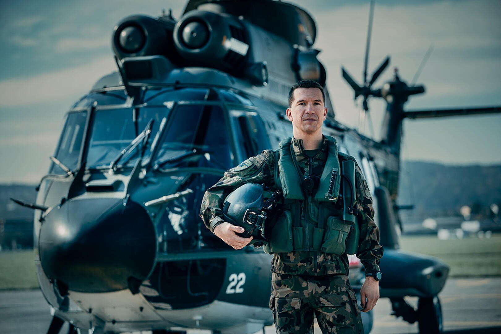 Ein Militärpilot in Tarnuniform und Ausrüstung steht selbstbewusst mit einem Helm vor einem dunkelgrünen Militärhubschrauber auf einem Flugplatz.