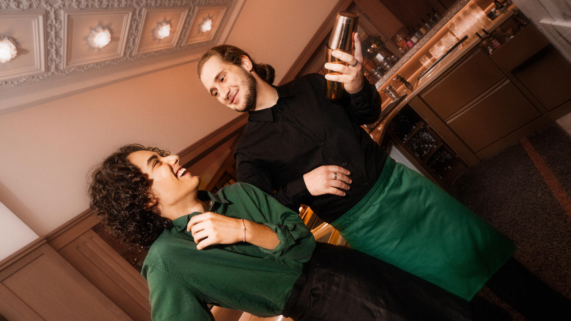 Zwei lächelnde Restaurantangestellte in grün-schwarzen Uniformen stehen zusammen; der eine schüttelt einen Cocktailshaker, während der andere lacht, beide wirken freundlich und engagiert in einer warm beleuchteten Bar.