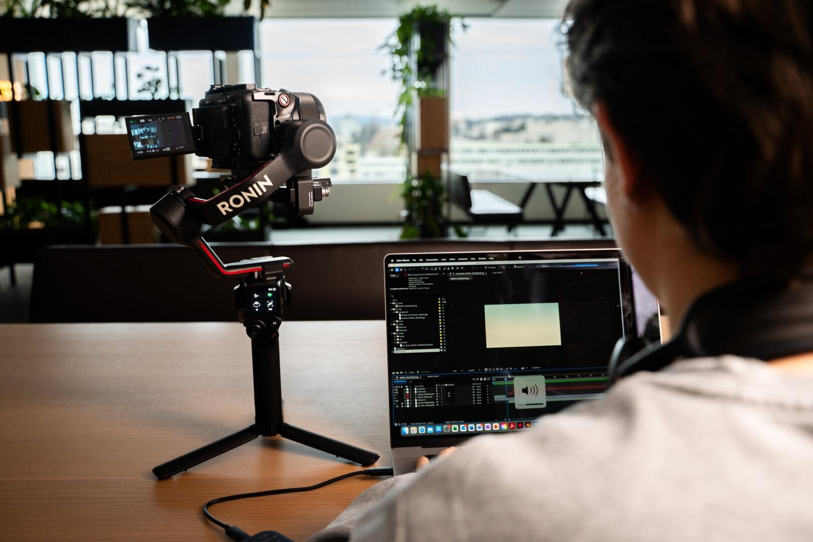 Eine Person arbeitet an einem Laptop mit geöffneter Videobearbeitungssoftware, während in der Nähe in einem modernen, gut beleuchteten Innenraum eine Digitalkamera auf einem kardanischen Ronin-Stativ montiert ist.